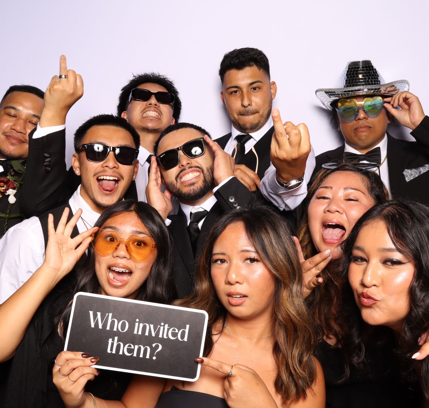 Group of friends posing in a photo booth, wearing black outfits, holding a sign that reads Who invited them.