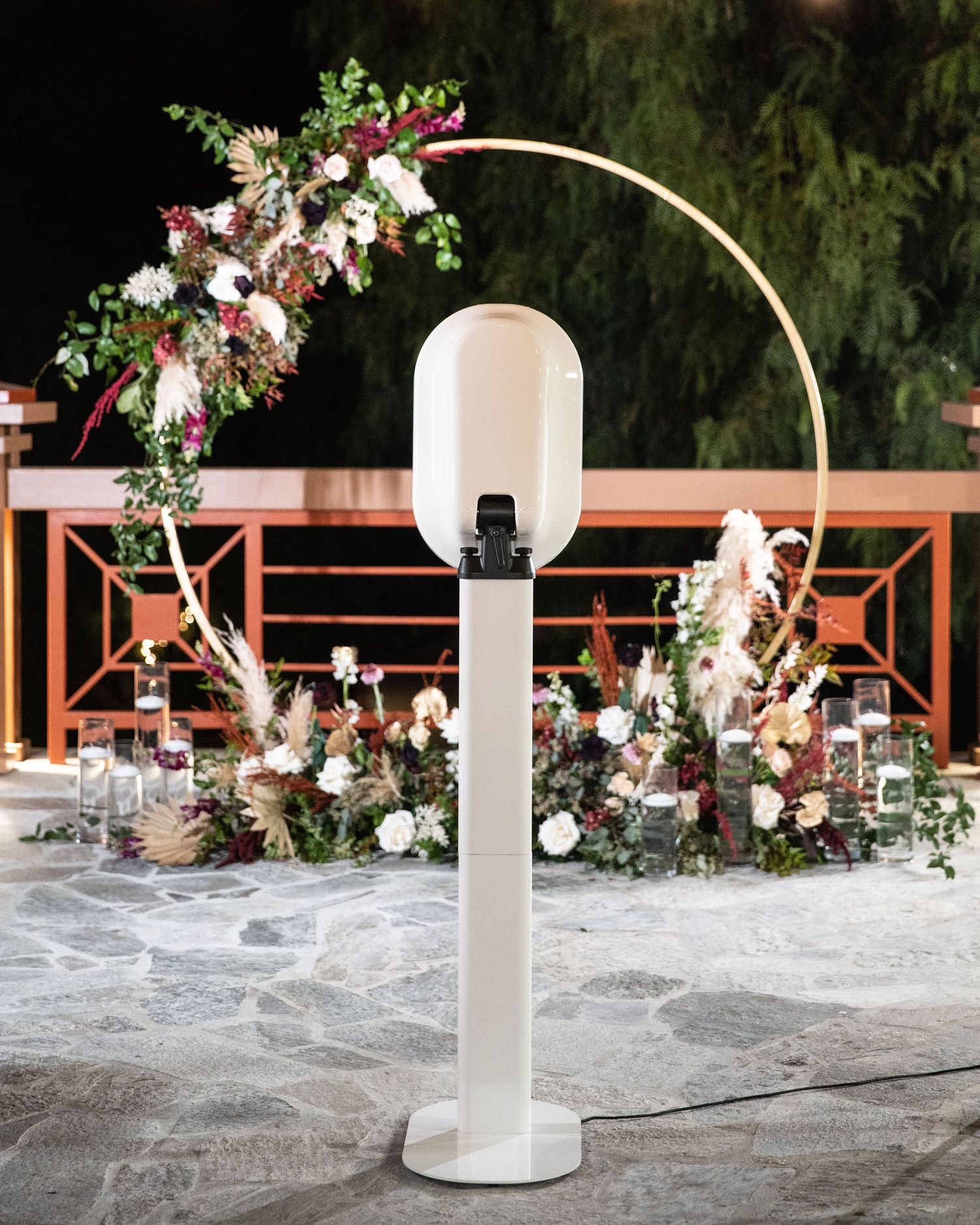 Elegant outdoor wedding setup with floral arch, white lantern stand, and lush flower arrangements on stone patio at night.