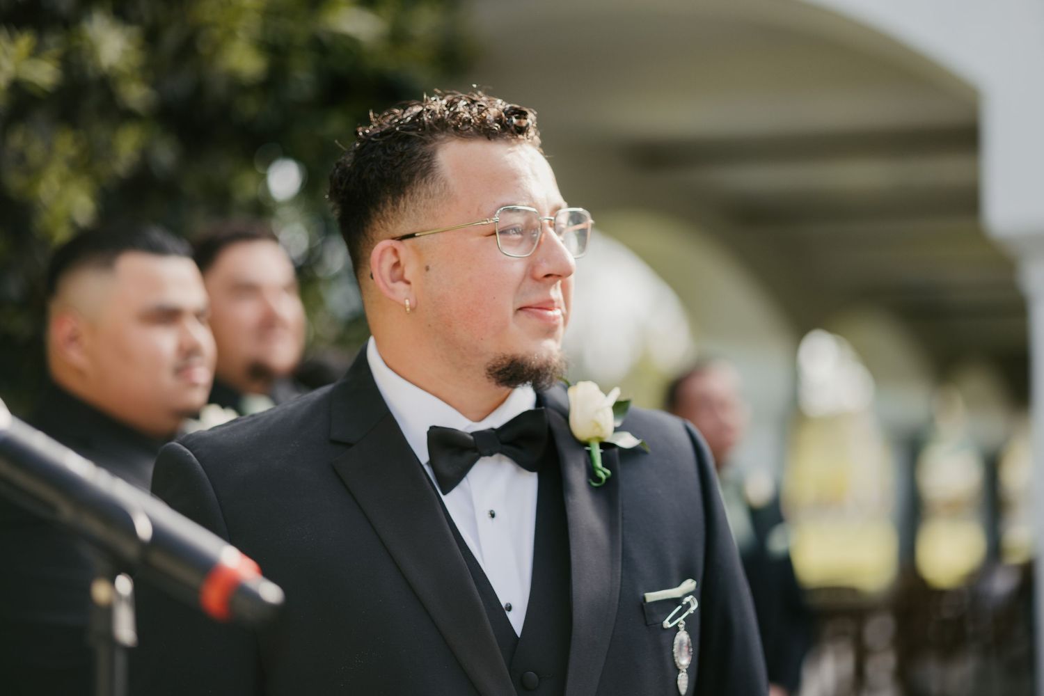 A formal wedding party member in a black tuxedo and glasses stands during an outdoor ceremony.
