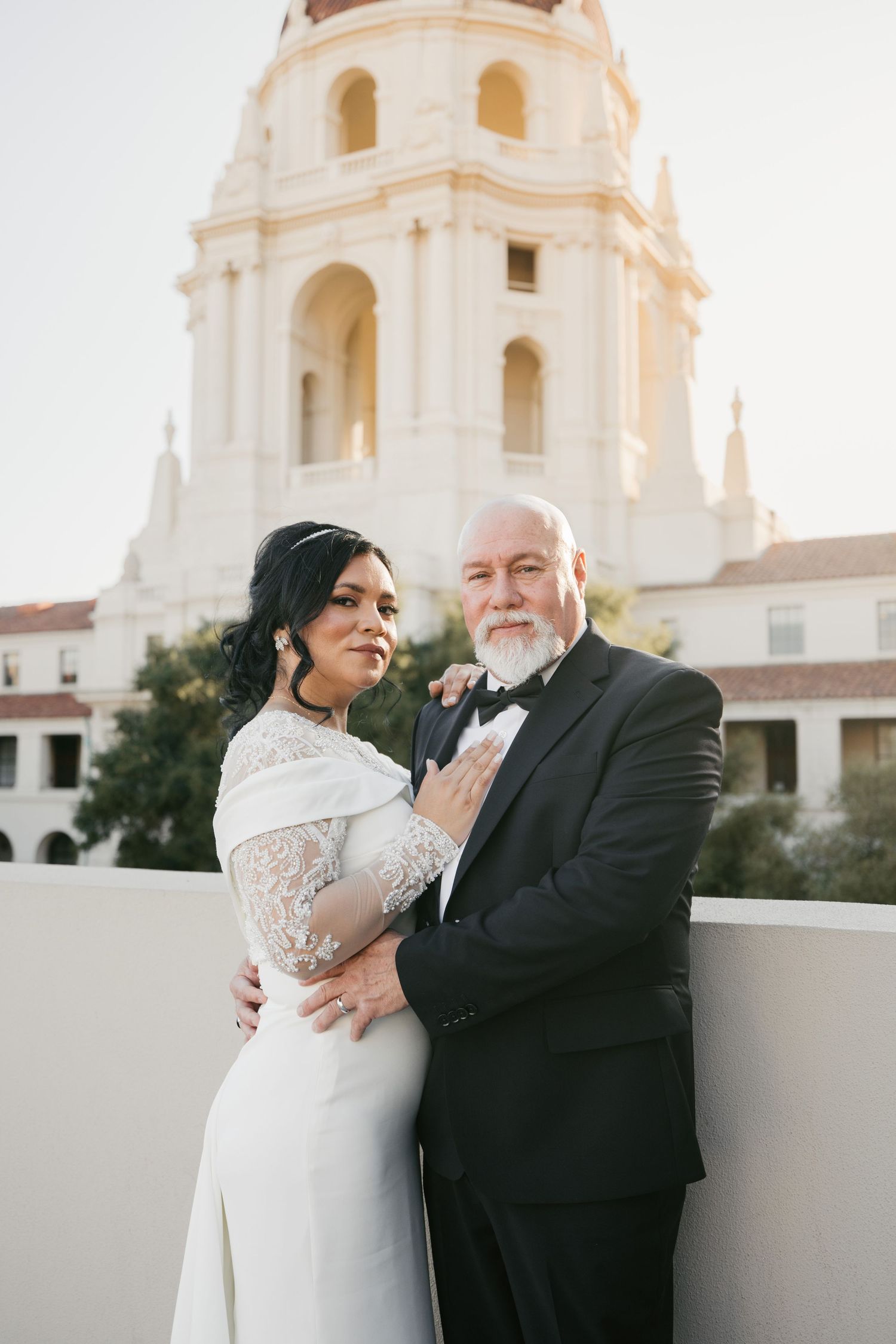 A couple poses together on a balcony with a Spanish mission tower in the background.