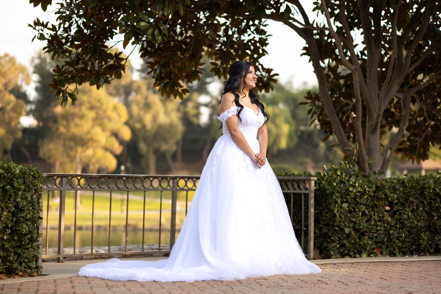 A bride in a white ballgown poses elegantly by a garden railing at sunset.