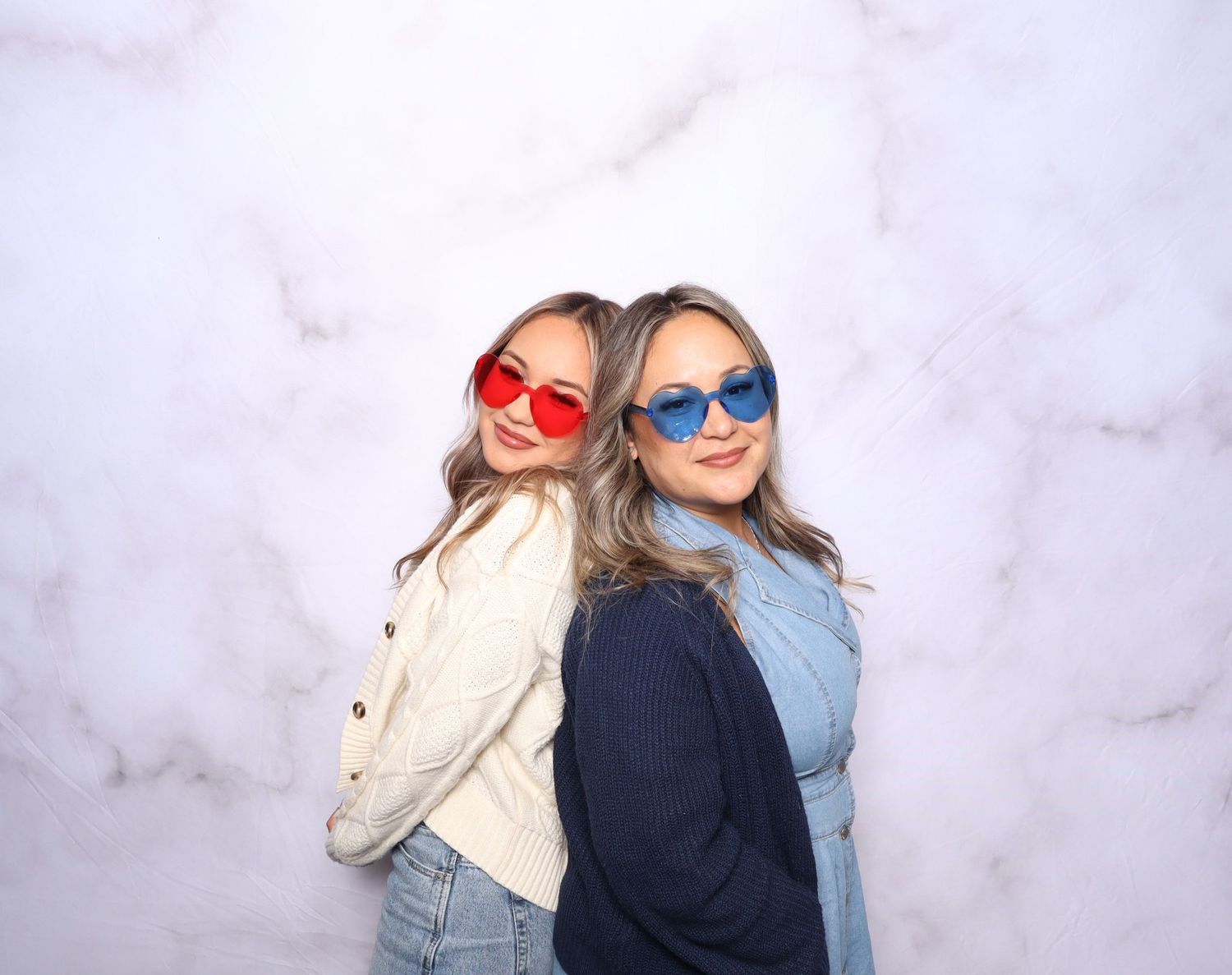 Two women wearing oversized red and blue sunglasses pose back-to-back against a marble background.