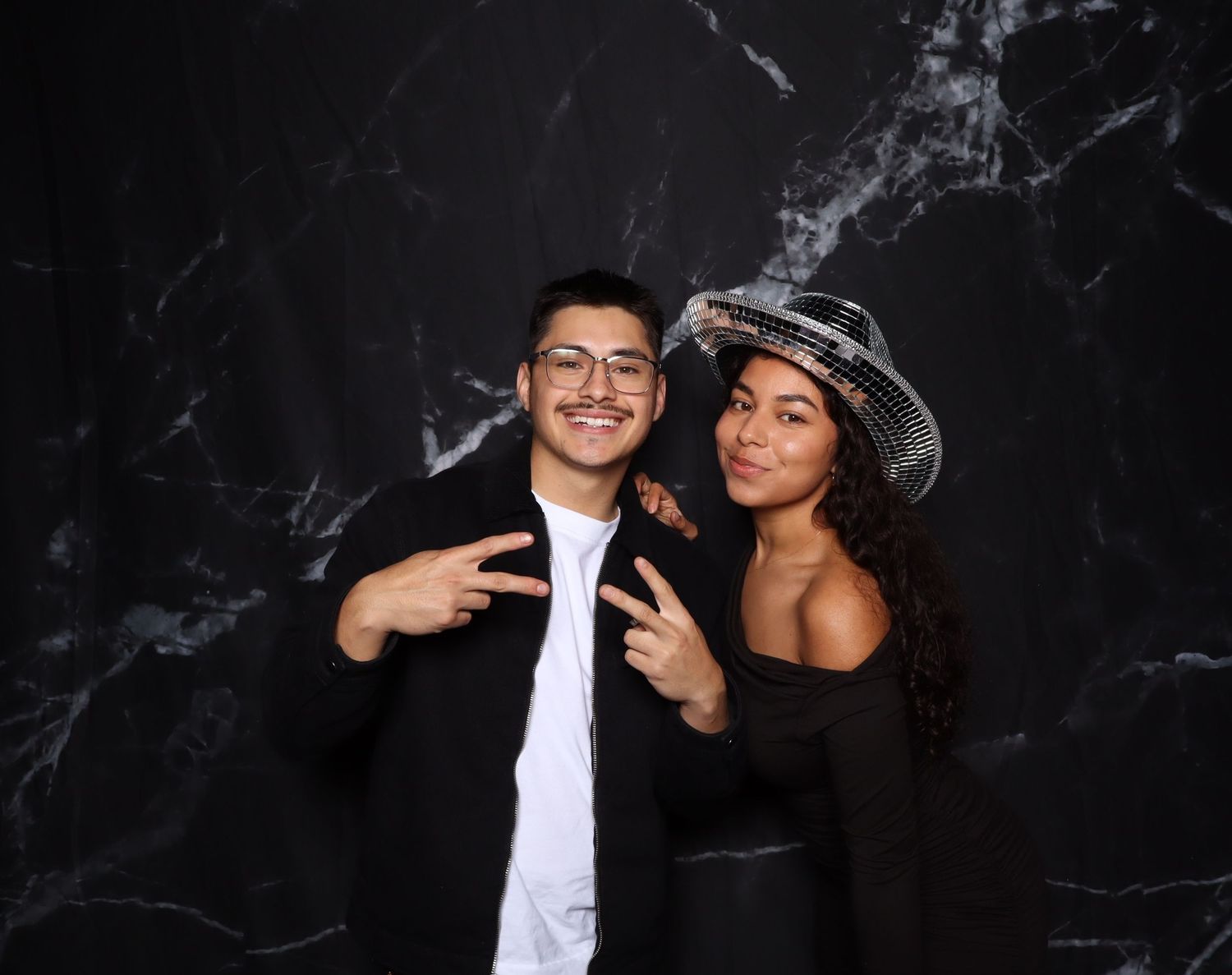 Two friends posing at a photo booth with marble backdrop, one wearing a sombrero hat and black dress.
