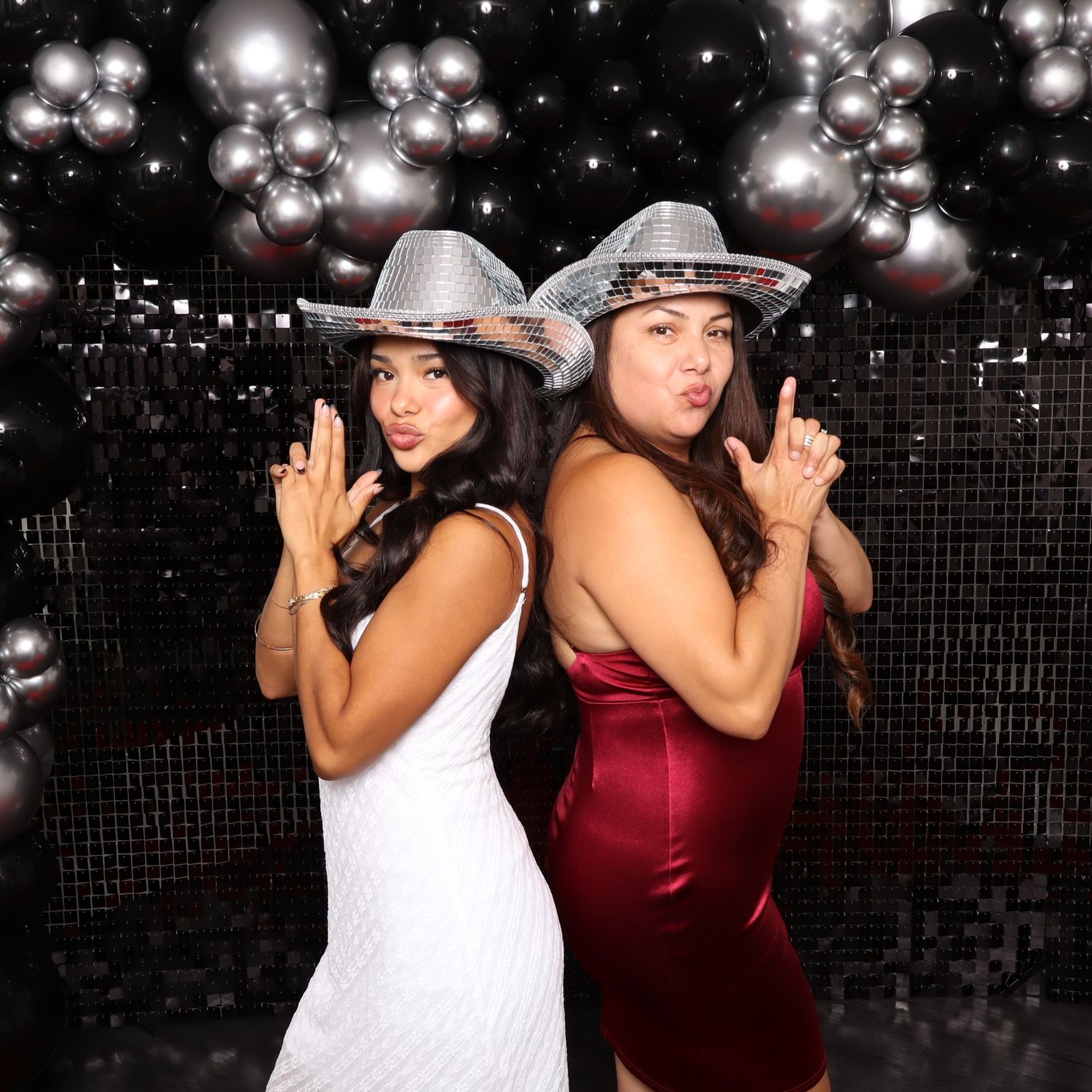 Two women in glam dresses and silver cowboy hats pose back-to-back at a party photo booth with balloon decor.