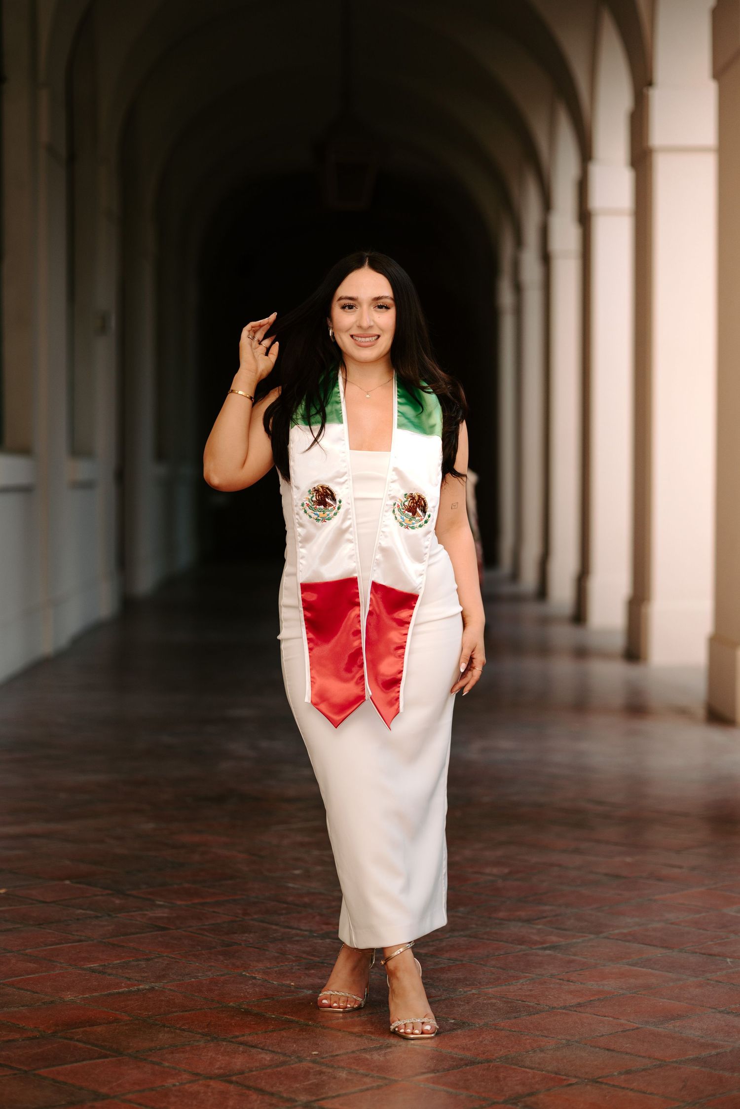 Graduate wearing a white dress with Mexican-themed decorative sash in a historic colonnade.