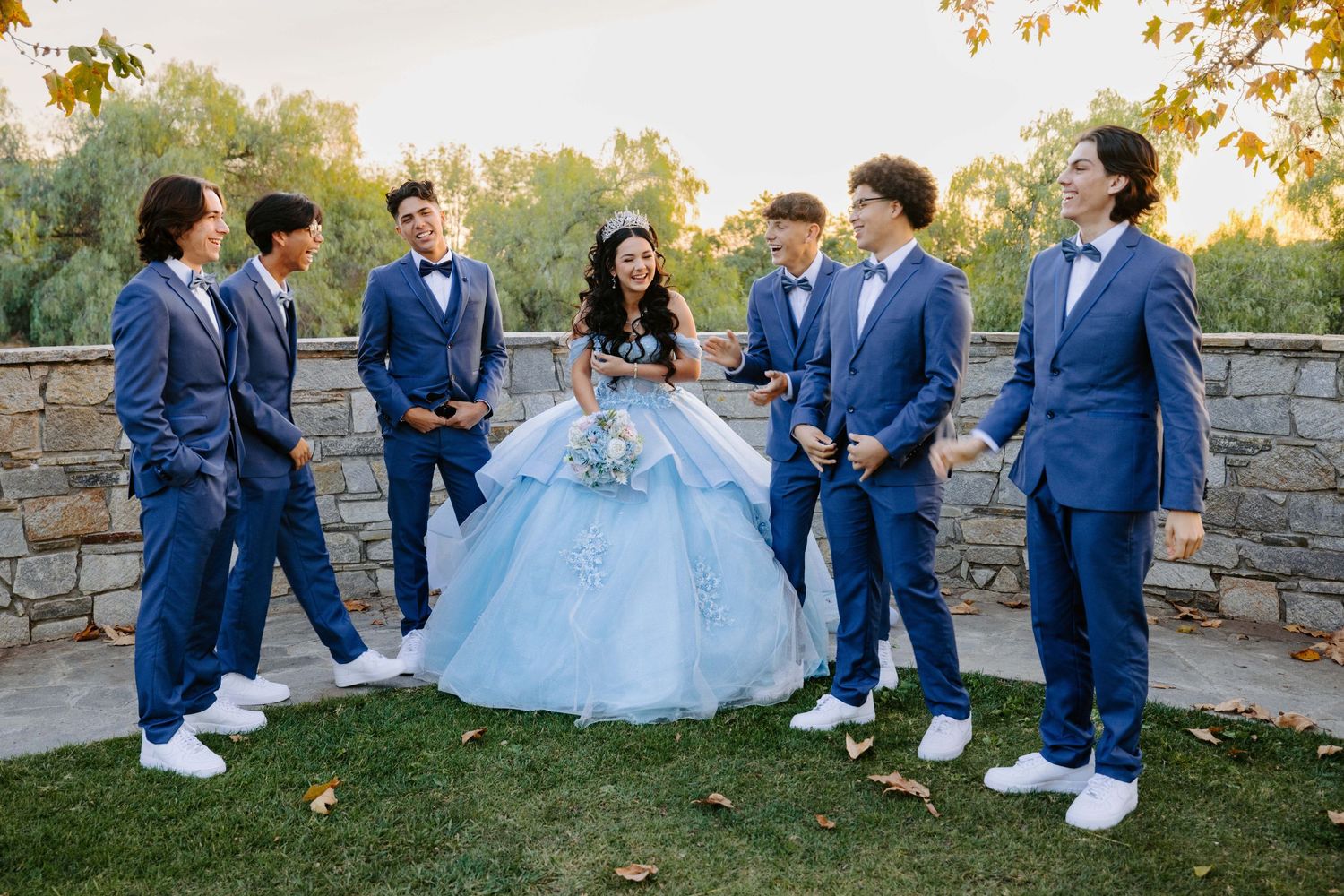 Group photo outdoors with people in coordinated navy suits and a light blue gown against a stone wall.