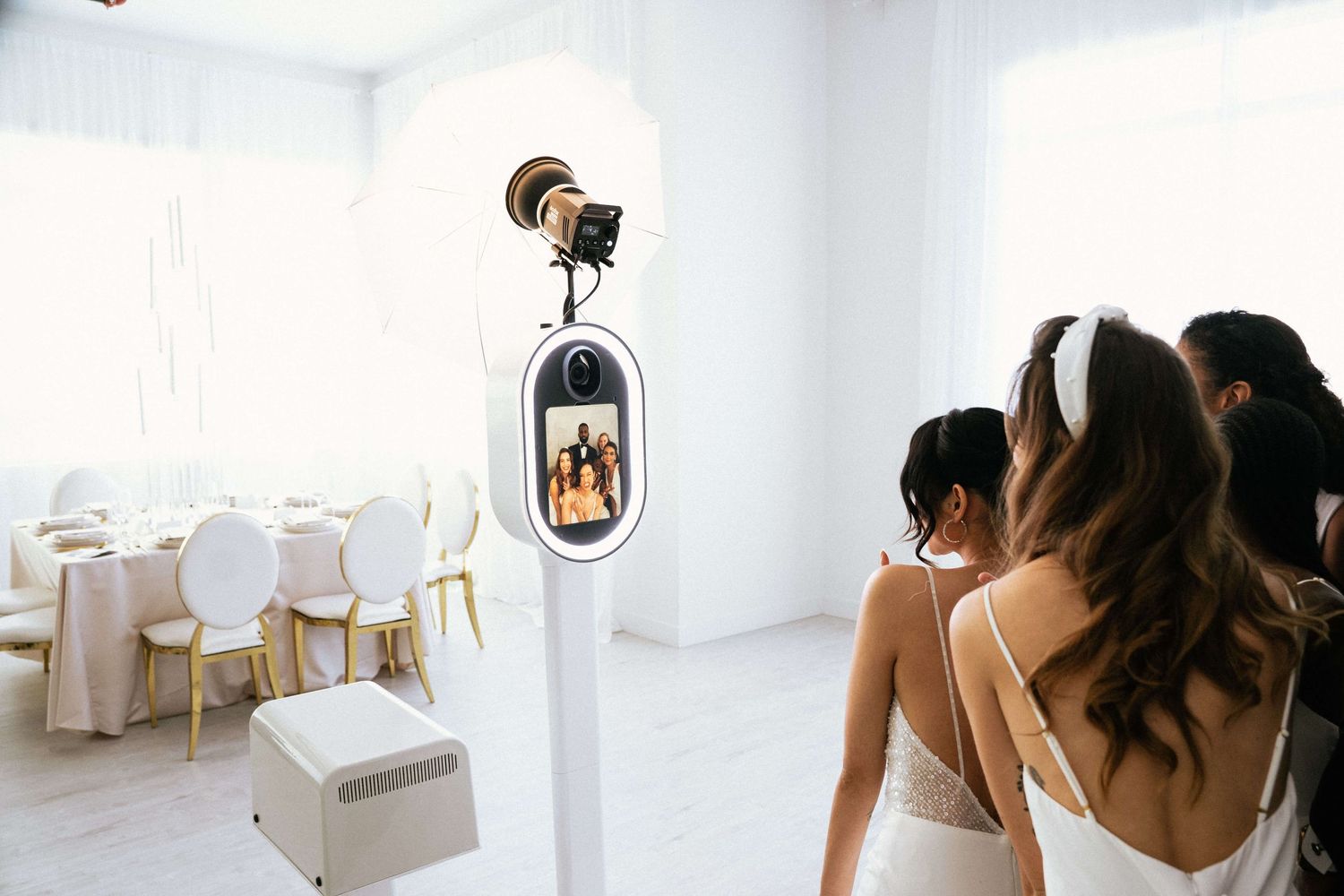 Brides posing at a modern photo booth kiosk in an elegant white wedding reception venue with gold chairs.