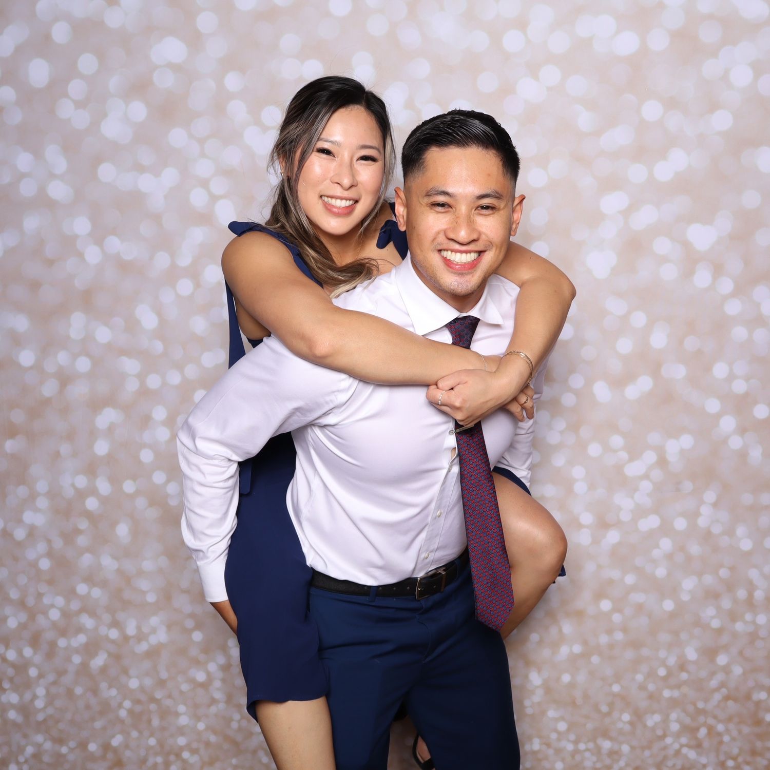 A smiling couple poses at a photo booth, man in white shirt and tie giving woman a piggyback ride.