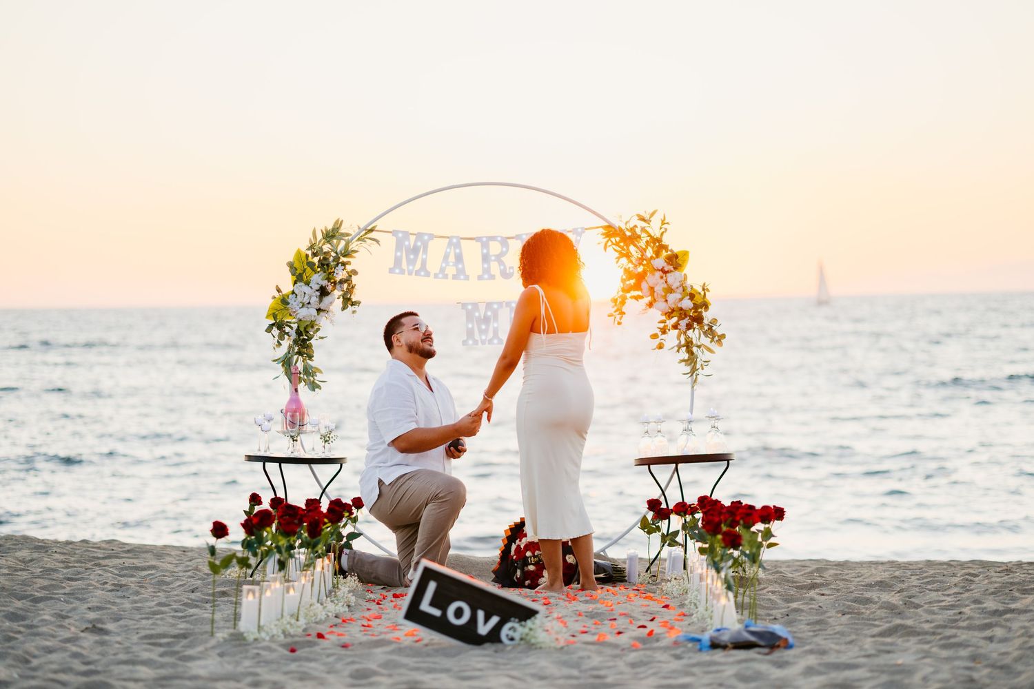 A sunset beach proposal scene with an illuminated floral arch and rose petal pathway by the ocean.