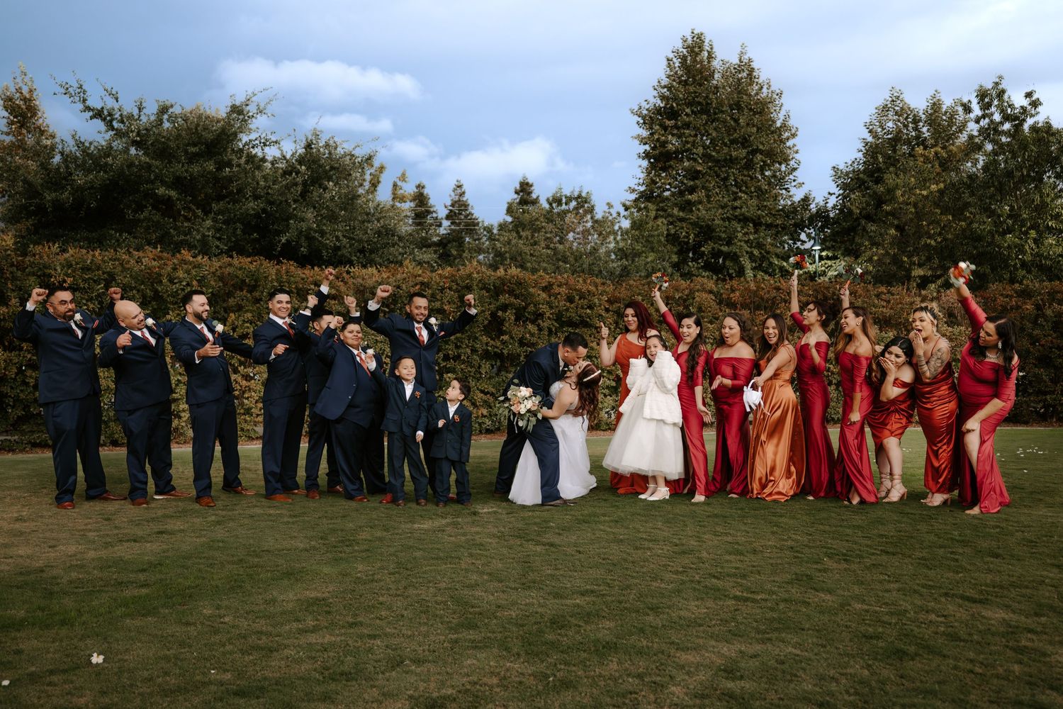 Wedding party poses outdoors on a sunny day with red and navy attire against a natural backdrop.