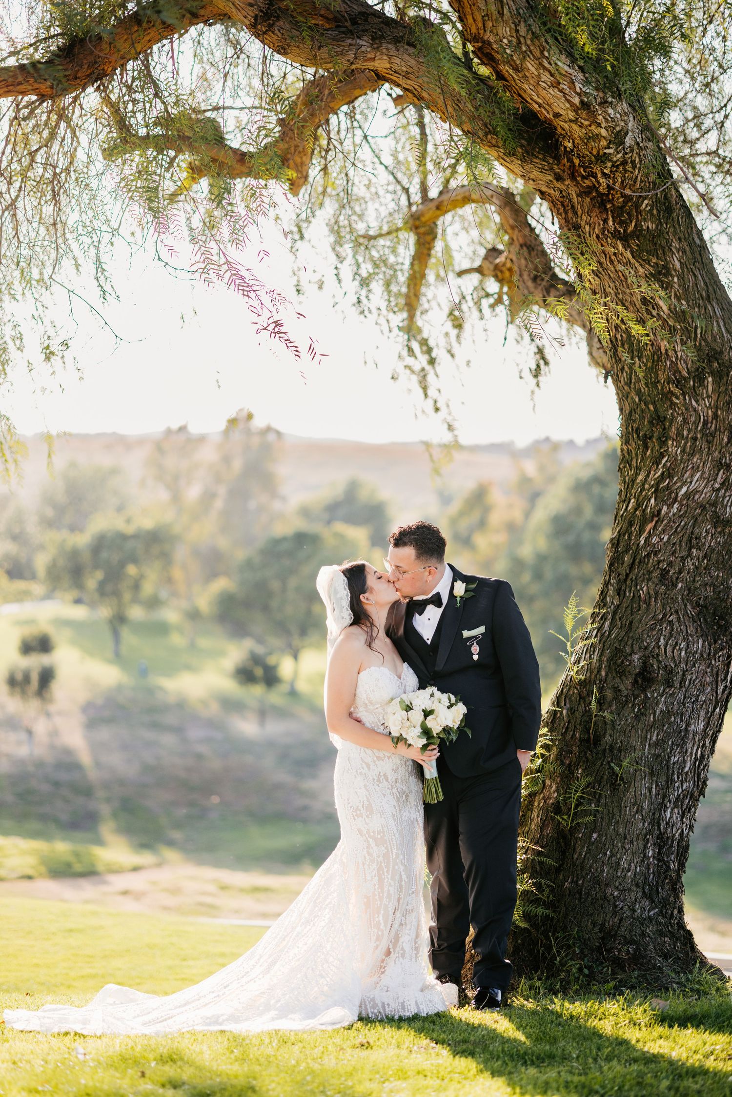A romantic wedding portrait under a large willow tree during golden hour.
