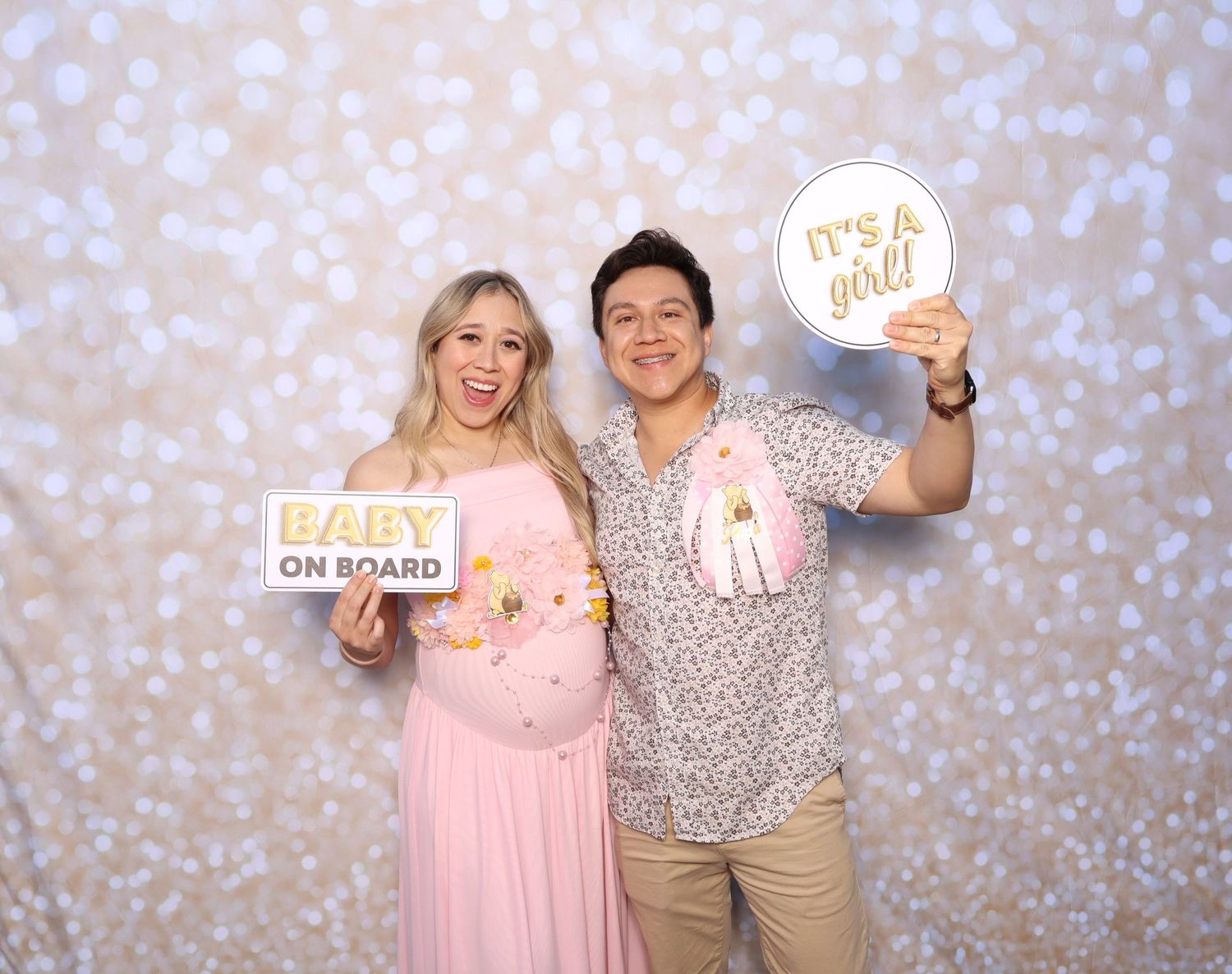 Couple at baby shower photo booth holding Baby On Board and It's a Girl signs in front of sparkle backdrop.