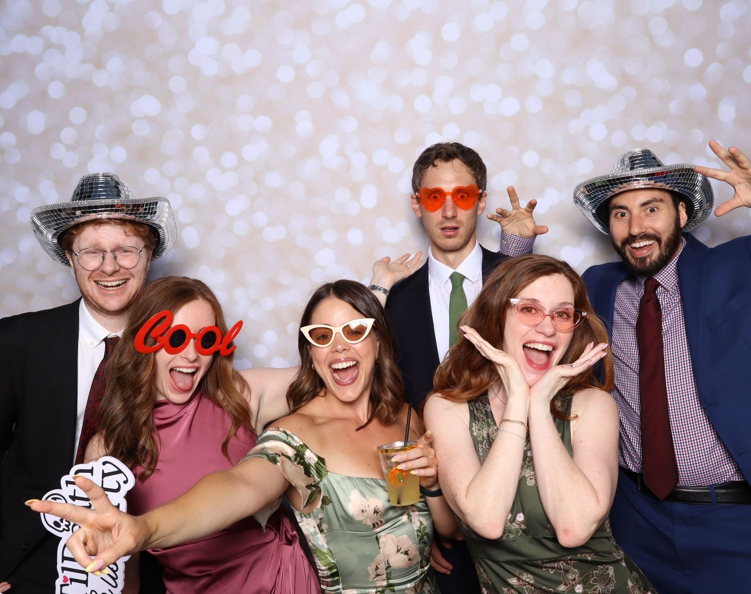 Six friends pose playfully in a photo booth wearing fun accessories like cowboy hats and colorful sunglasses at a wedding reception.