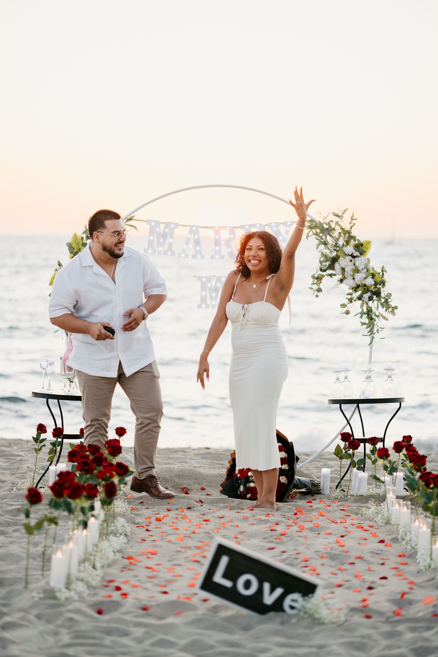 A couple celebrates joyfully under a decorated arch on the beach with scattered rose petals and ocean views.