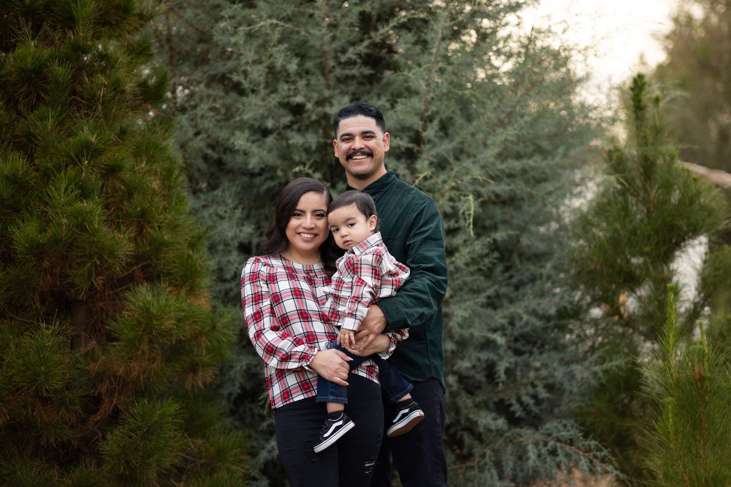 Family embracing outdoors with evergreen trees in background during portrait session.
