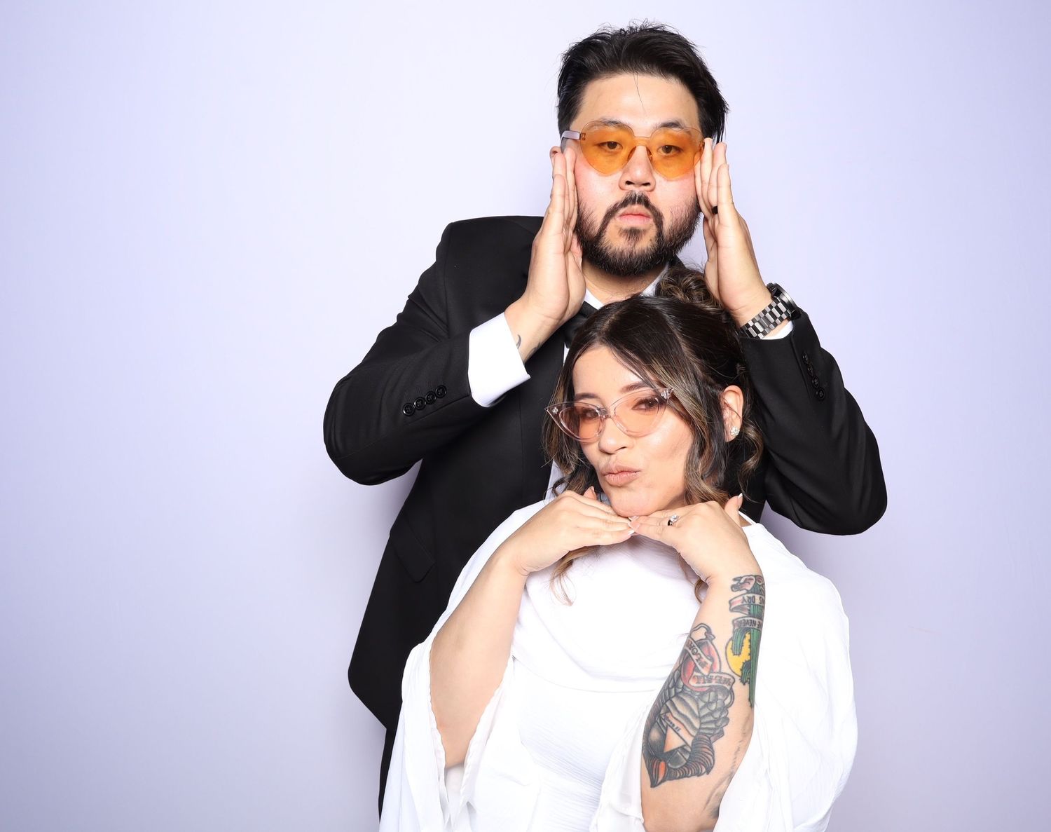 A bearded man in a black suit and a woman in white pose together, both wearing orange tinted sunglasses.