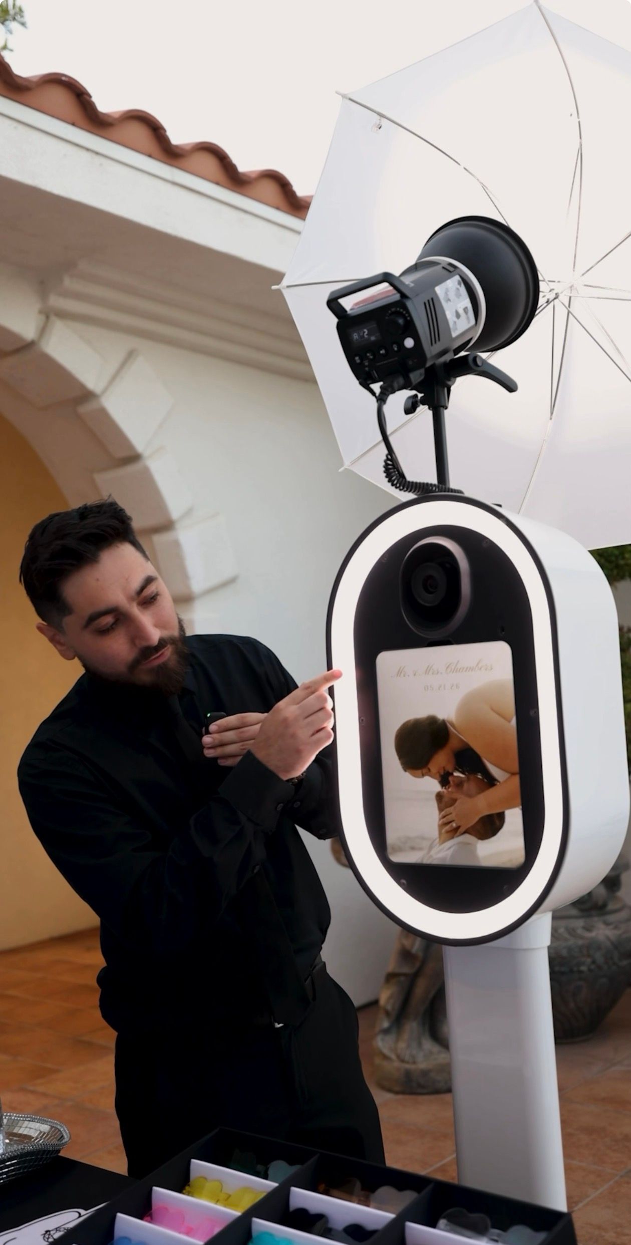 Man in black adjusting a modern photo booth with ring light and flash umbrella at an outdoor wedding event.