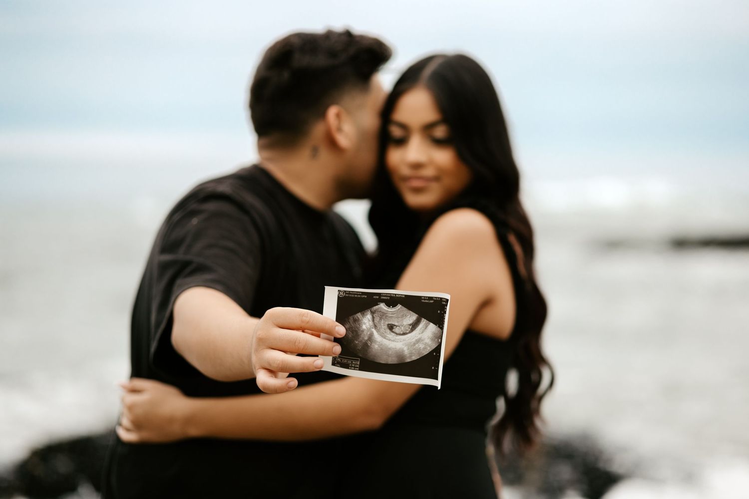 Intimate couple sharing moment on beach while holding ultrasound photograph.