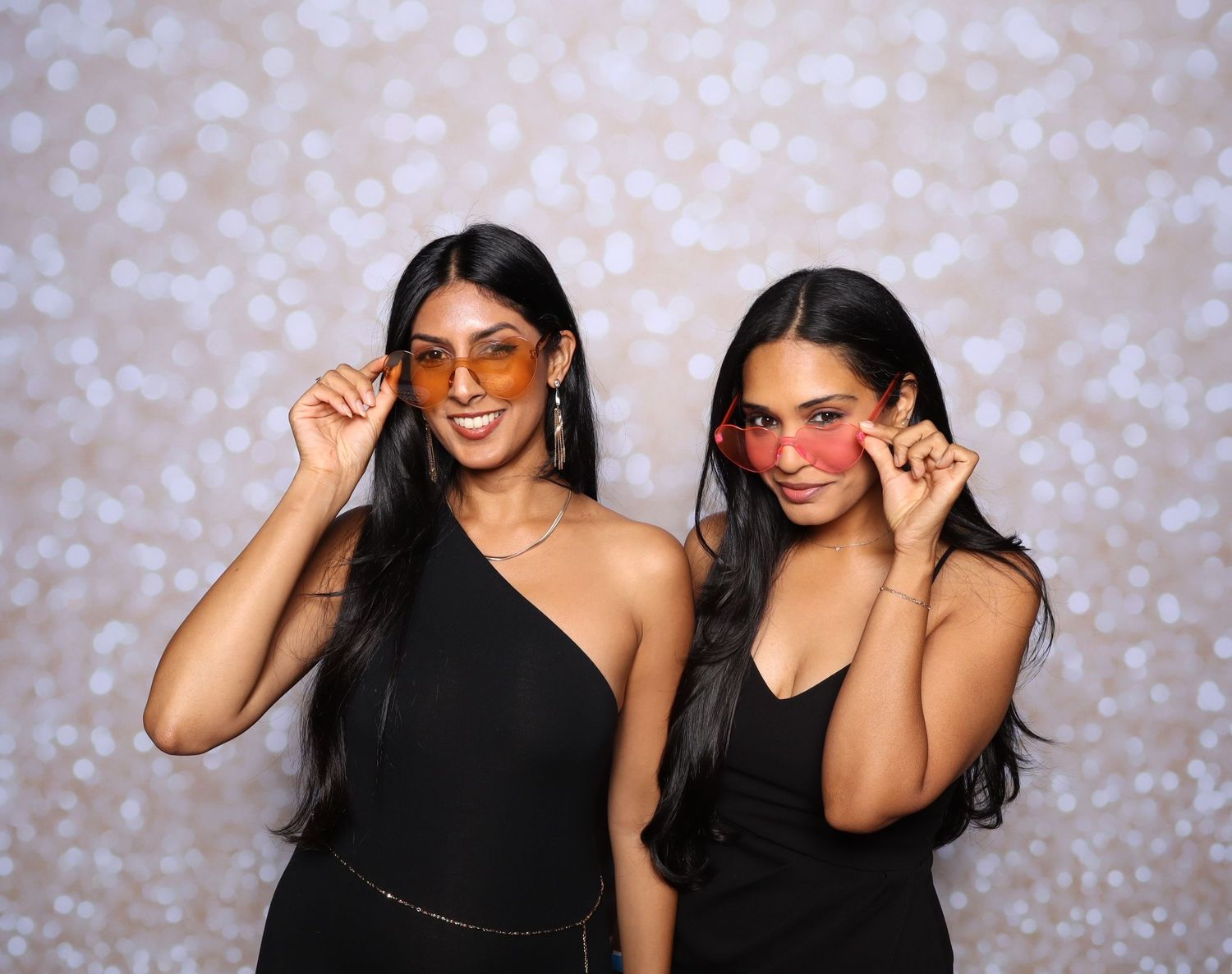 Two women in black outfits pose with colorful sunglasses against a bokeh backdrop.