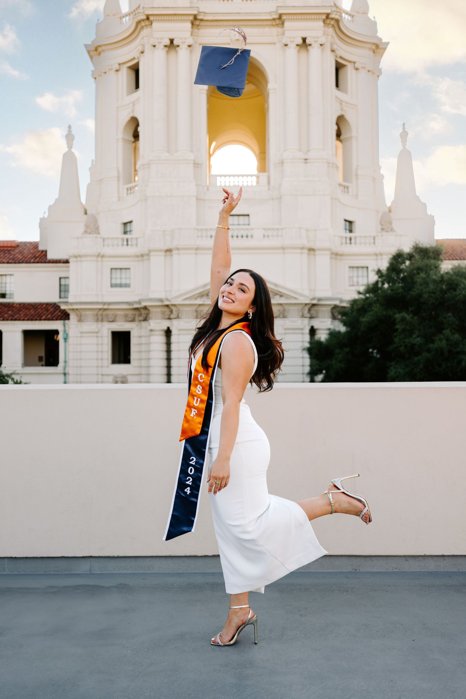 Graduate celebrating with diploma and sash in front of white church tower architecture.