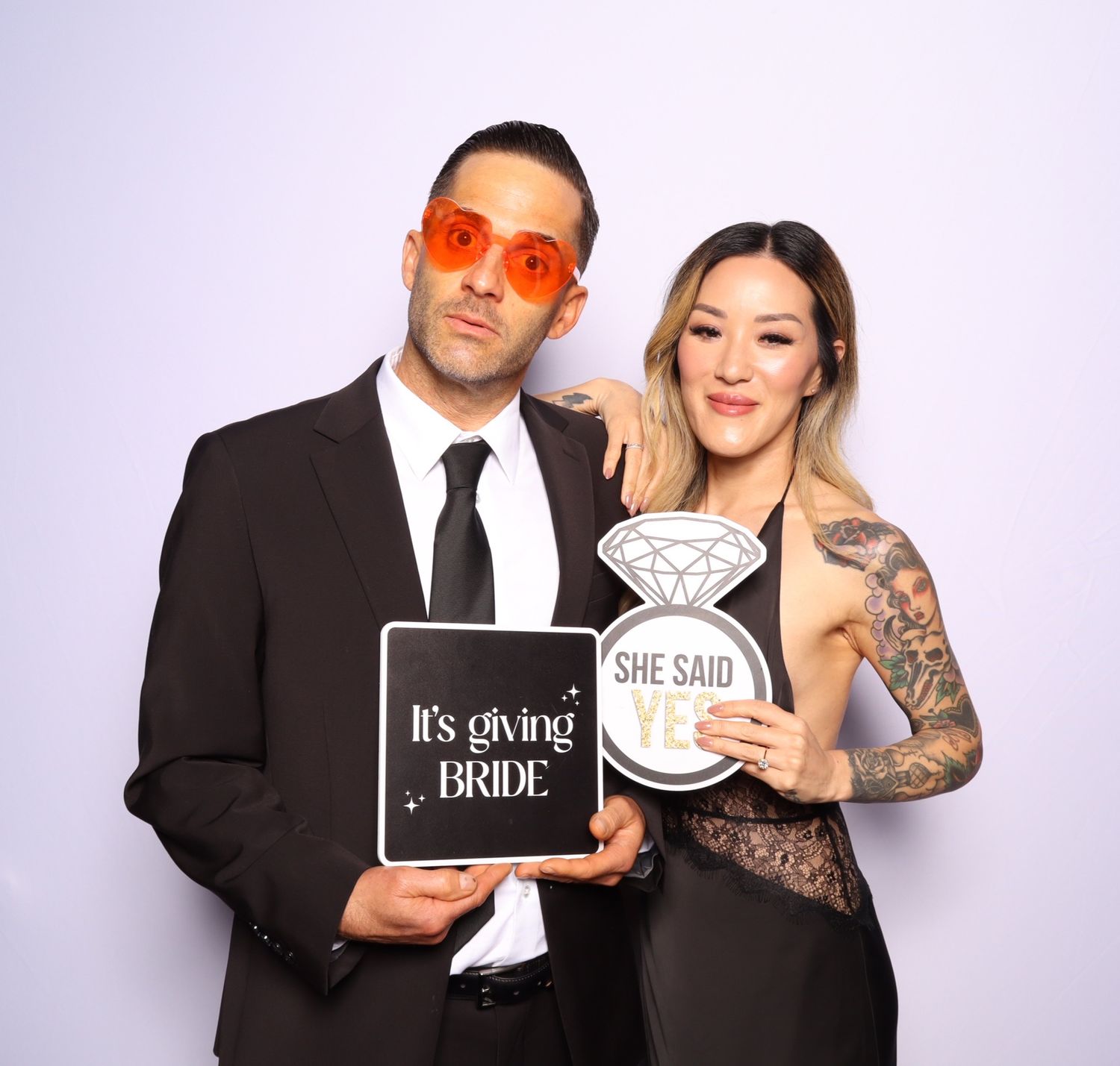 Couple posing in photo booth holding bride signs, man in suit with orange glasses, woman in black lace dress.