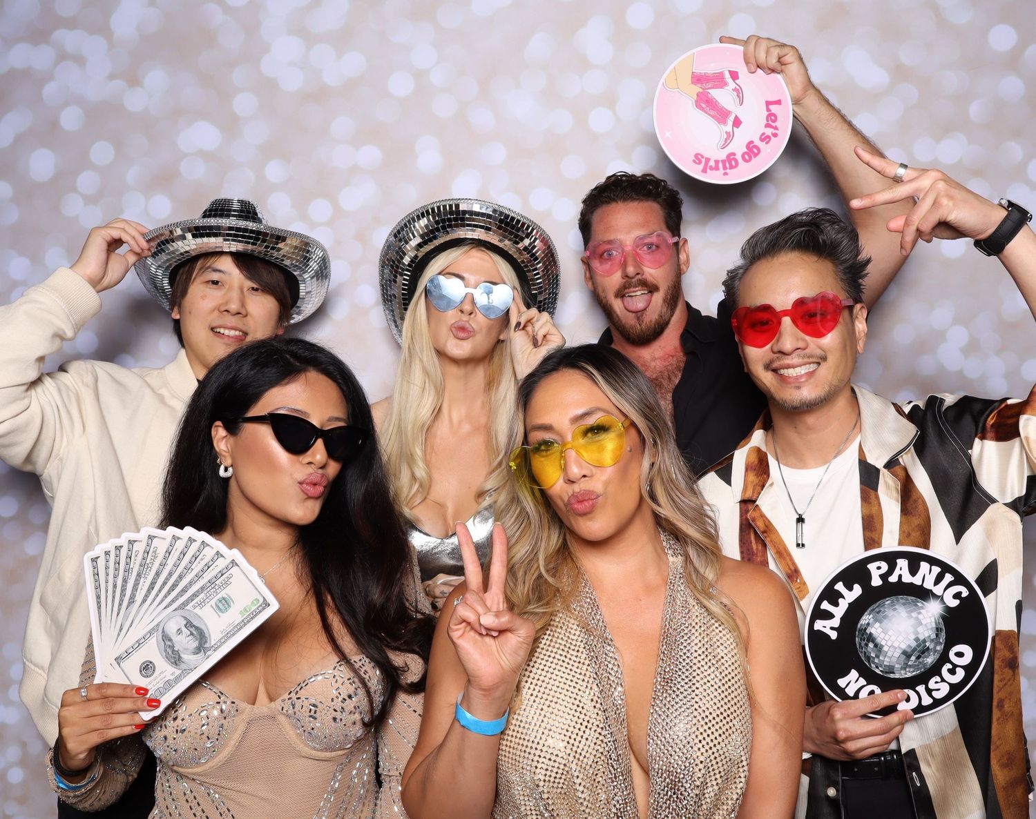 Group of people wearing sunglasses and festive attire pose together in a photo booth strip with silver bokeh background.