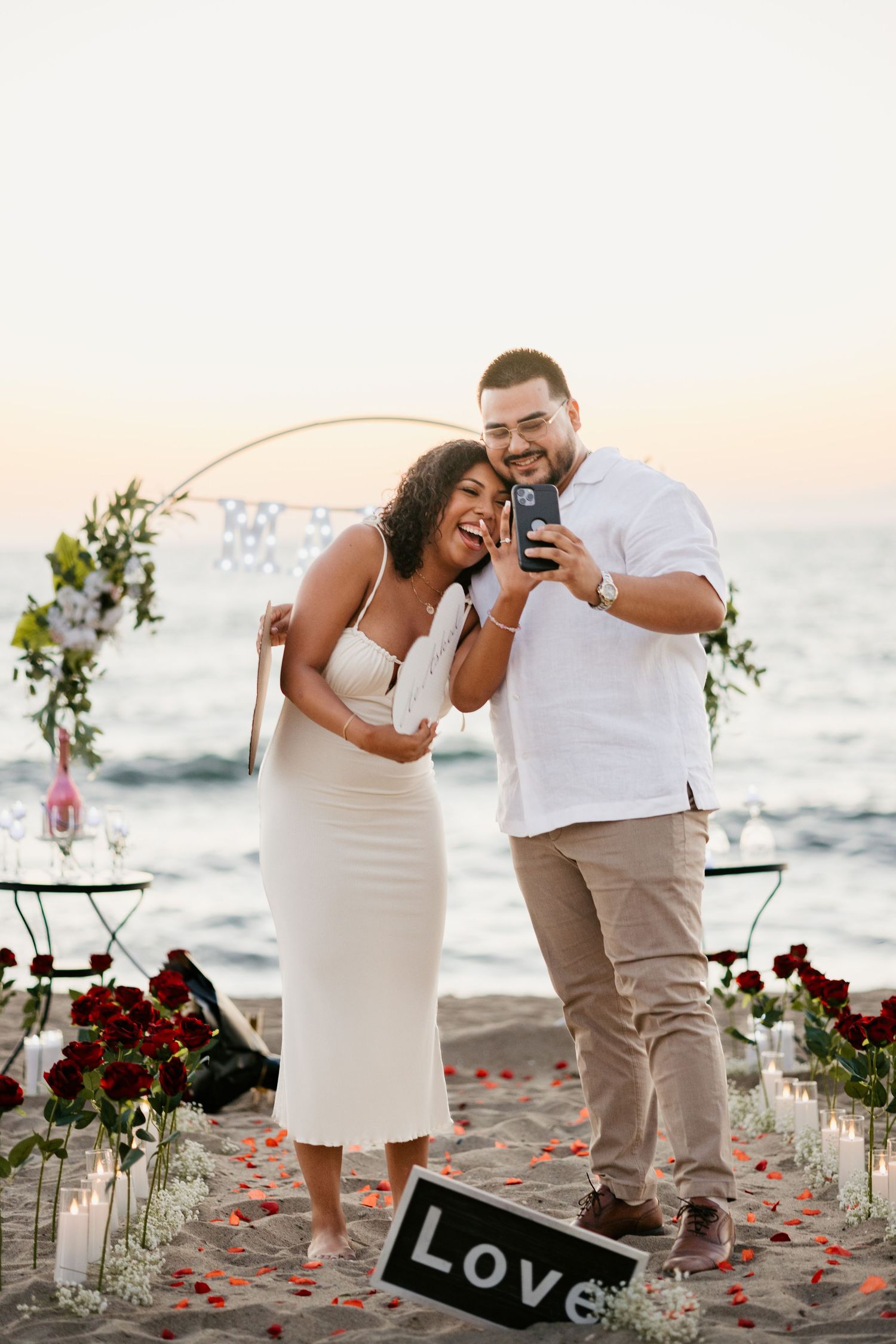 A couple shares a joyous moment on a beach with romantic decorations and a 'LOVE' sign.