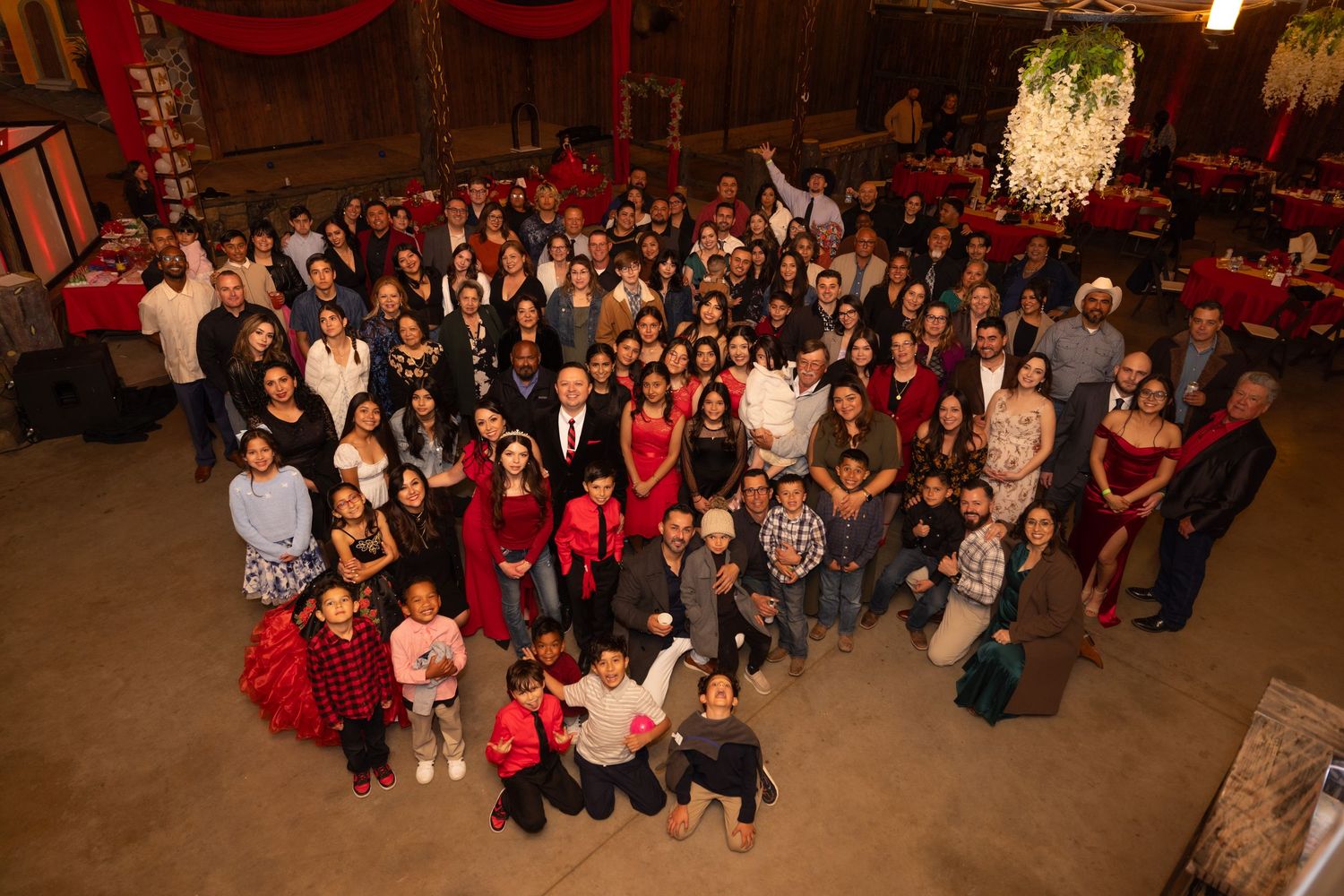 Large group celebration photo in formal attire inside rustic venue with red decor.