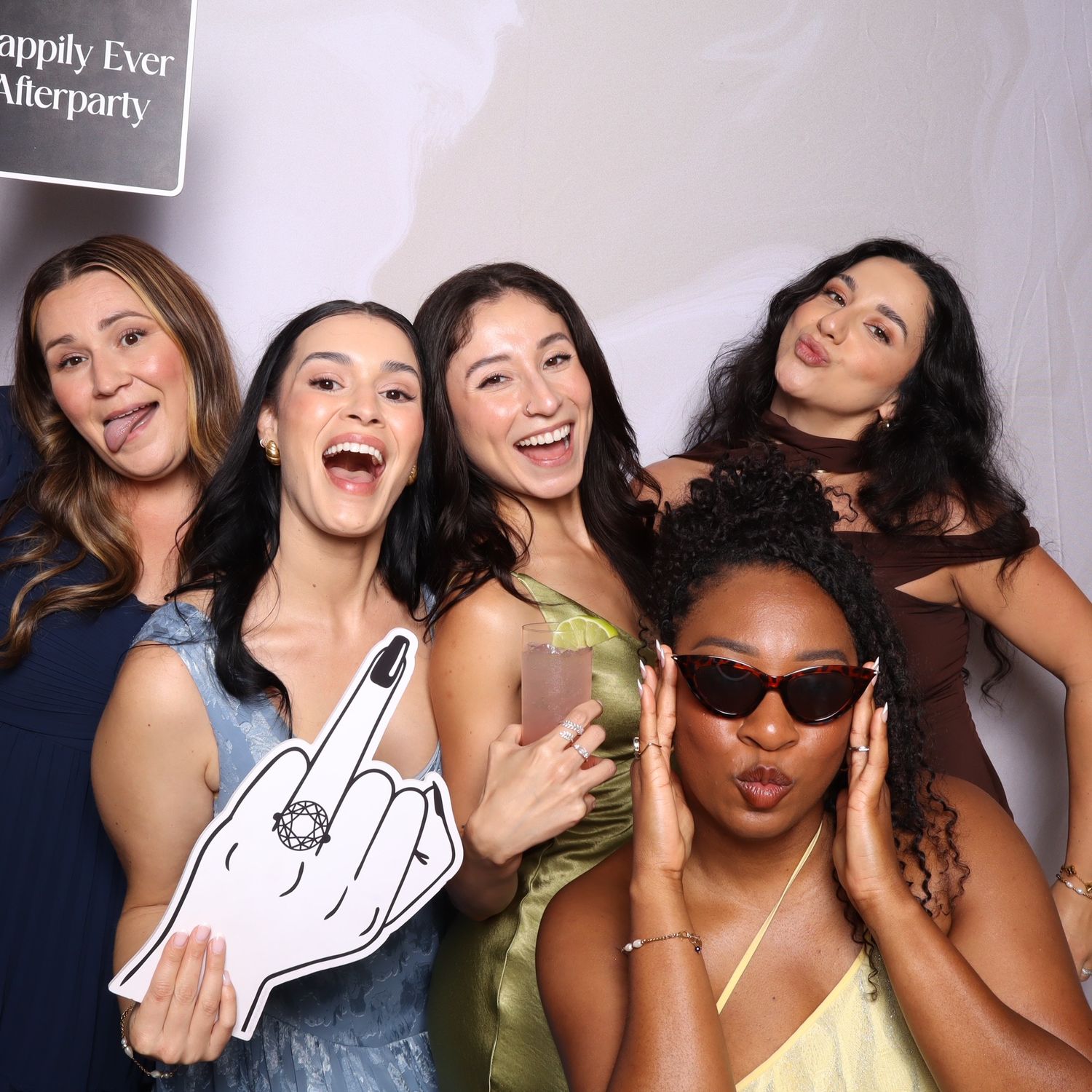 Five women posing playfully at a Happily Ever Afterparty photo booth, one holding a foam finger prop.