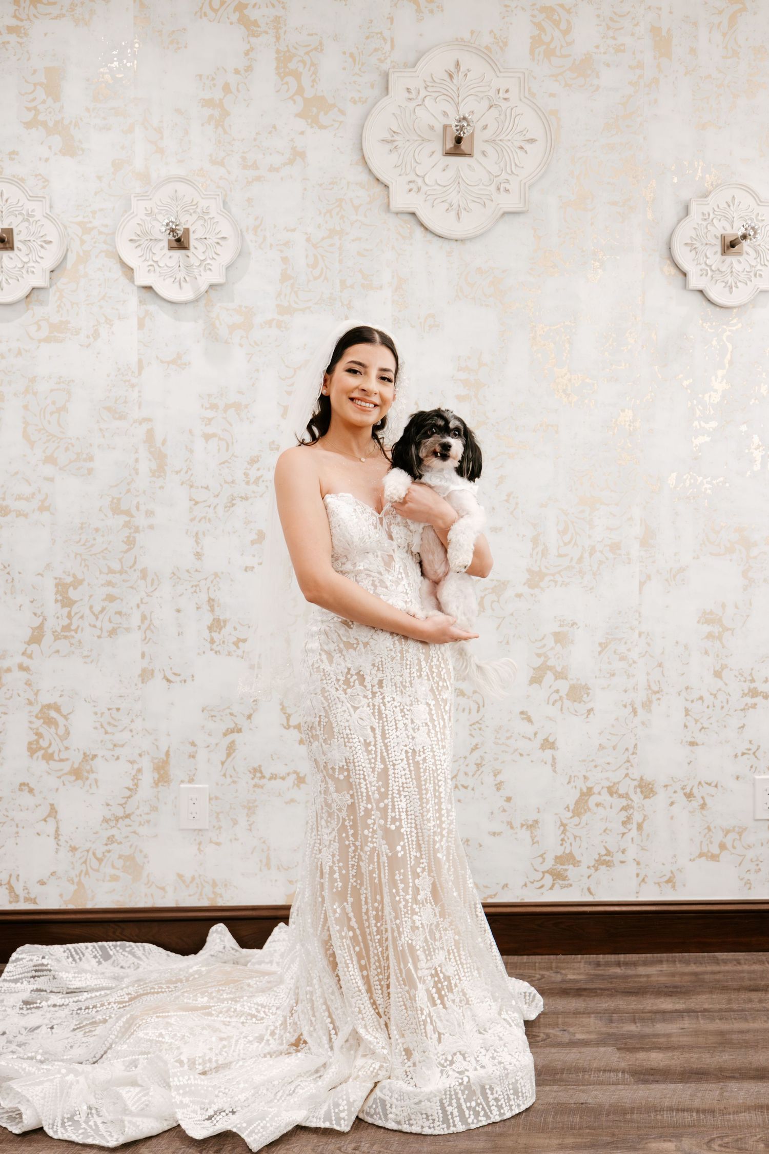 Bride in sparkly gown holds small black and white dog against floral wall backdrop.
