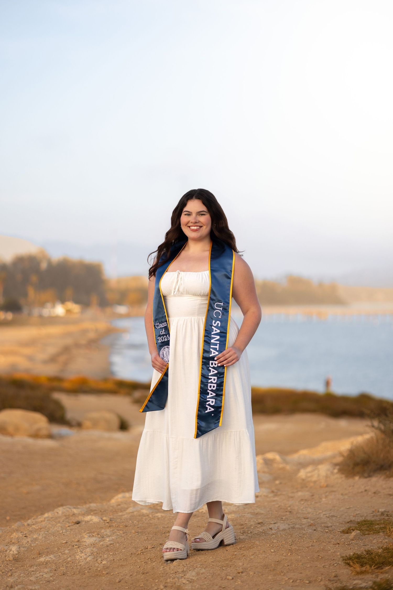 Graduate wearing white dress and blue graduation sash poses on rocky coastal landscape.
