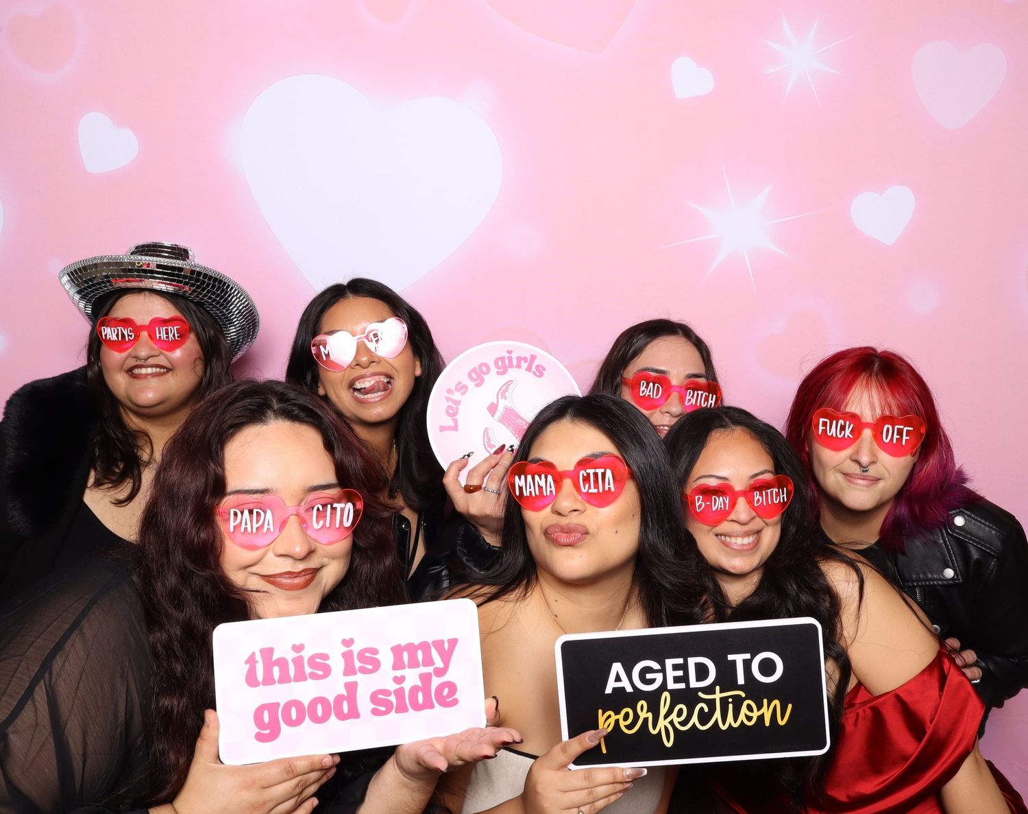 Group of seven women posing in photo booth with fun props and signs reading 'Aged to Perfection' and 'This is My Good Side'.