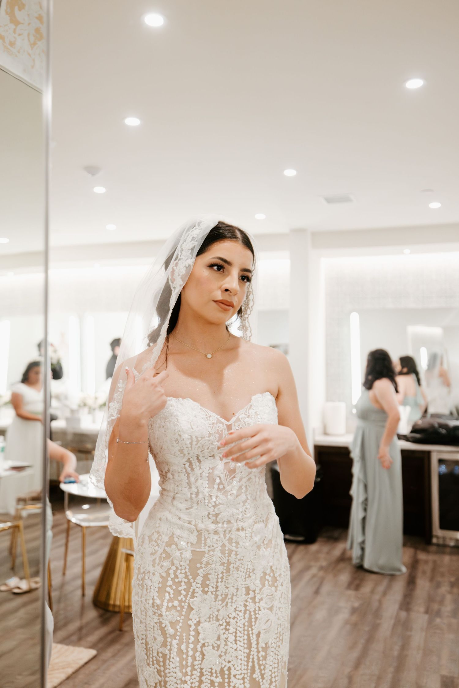 Bride in lace wedding gown standing in bridal suite with mirrors.