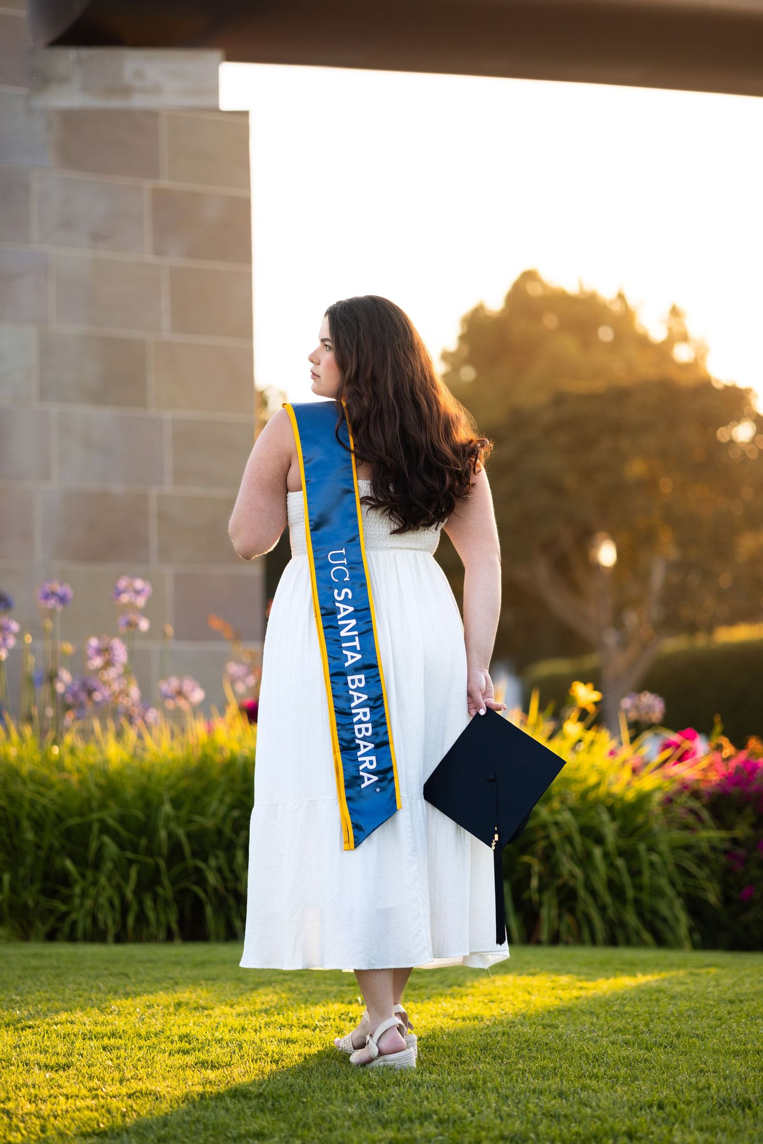 White graduation dress and ceremonial sash photographed against blooming garden landscape.