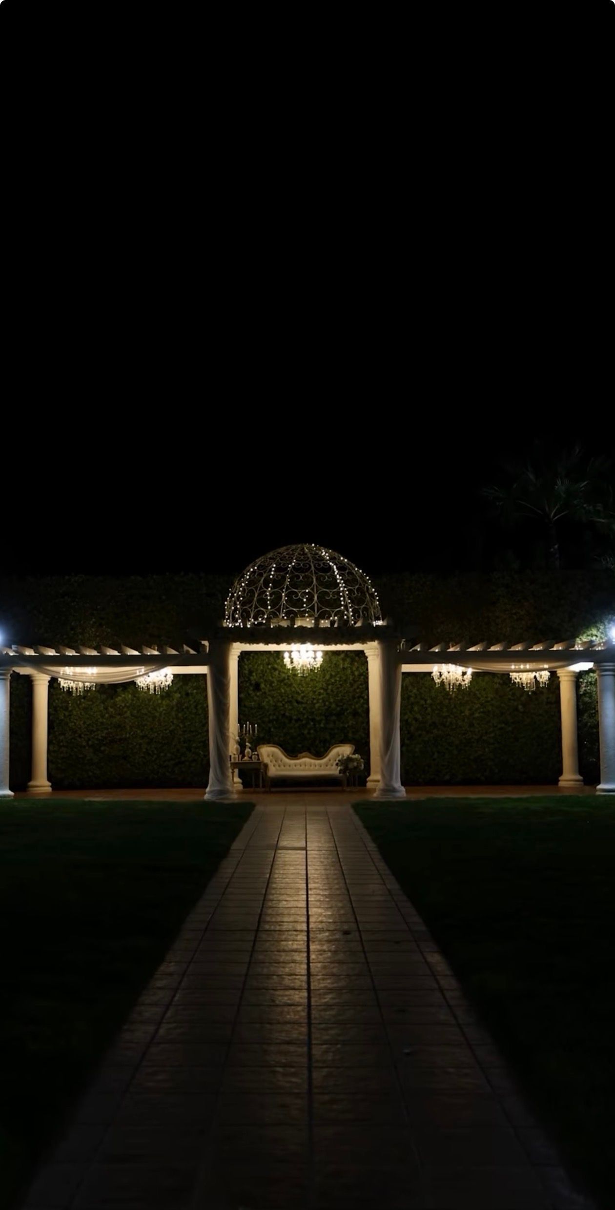 A lit gazebo with a domed roof and columns stands at the end of a paved pathway at night, creating a romantic ambiance.