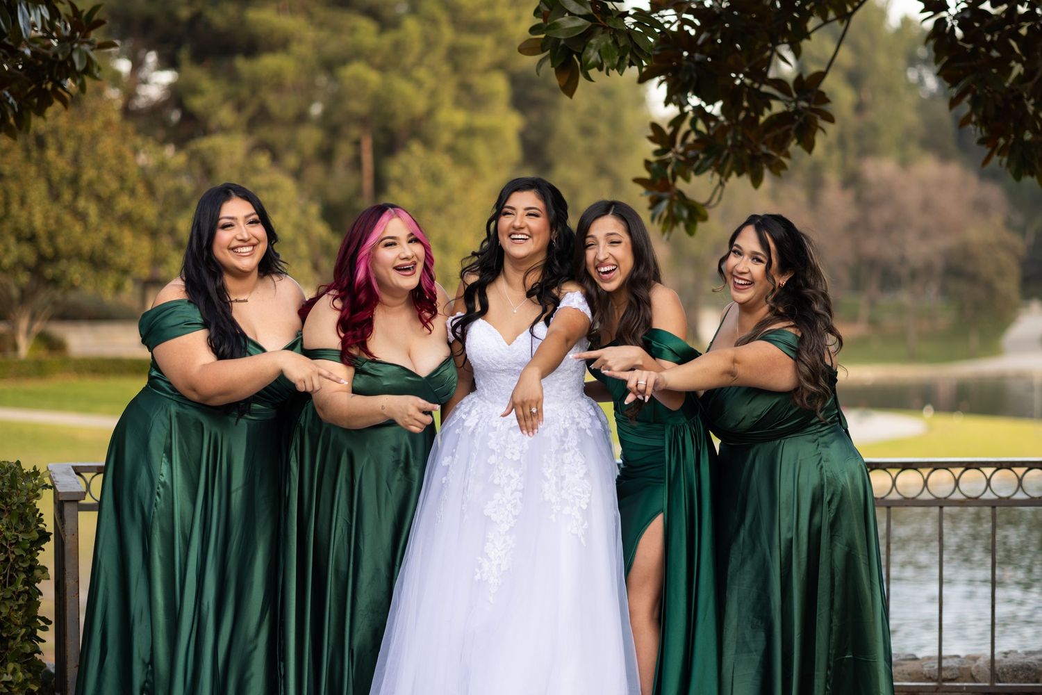 Bridesmaids in elegant emerald green dresses pose together outdoors near a lake.