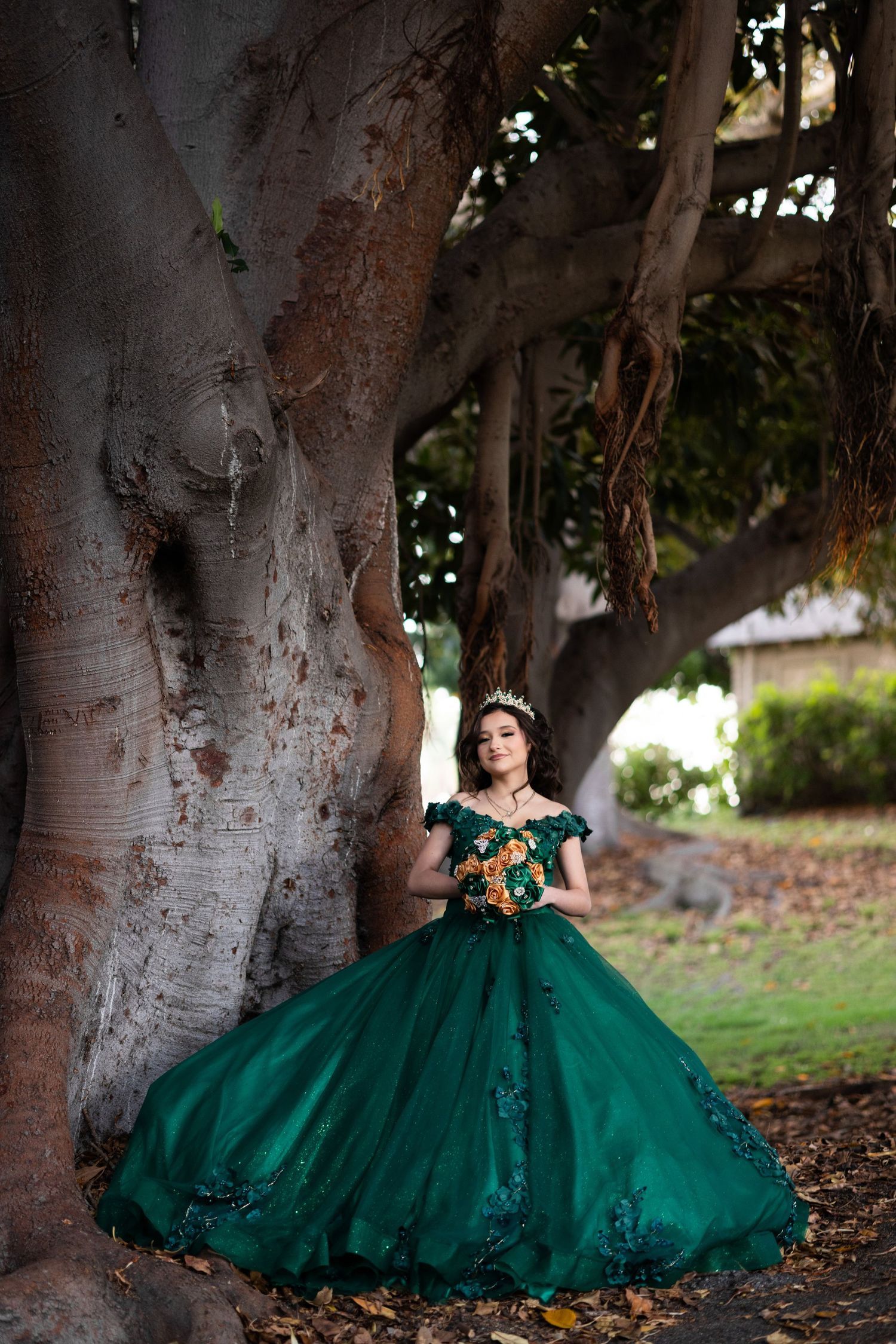 Stunning emerald green ball gown with intricate details photographed against large tree trunks in natural lighting.