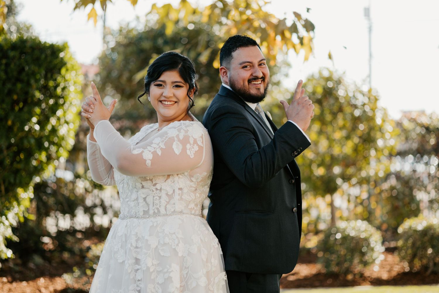 A wedding couple stands back-to-back outdoors with arms raised in a playful pose at sunset.