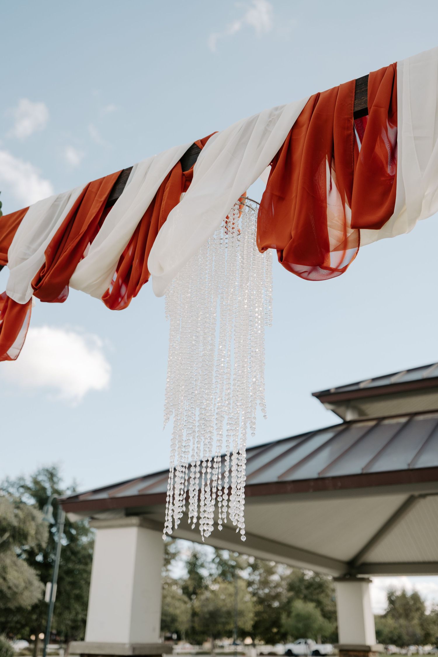 Red fabric and crystal decorations hang elegantly against a pale blue sky at an outdoor wedding venue.