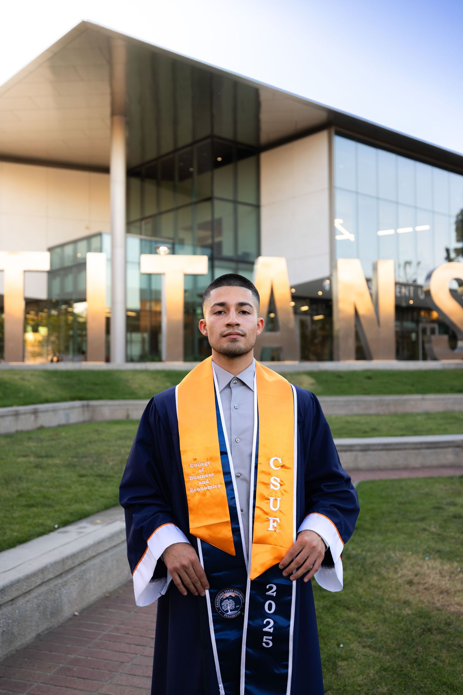 Graduate wearing navy blue cap and gown with gold sash poses outdoors in front of modern campus building.