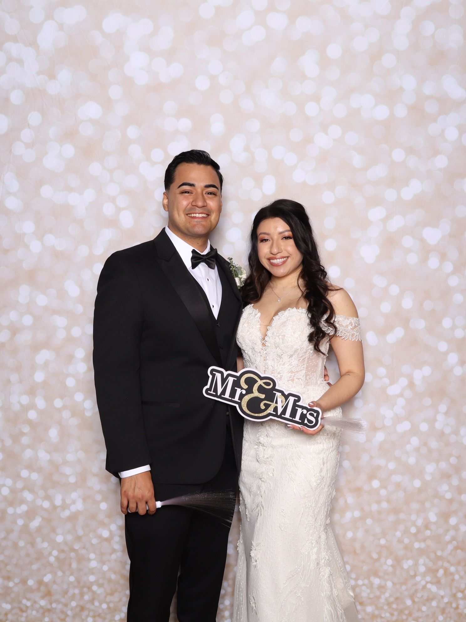 A bride and groom pose together at their wedding against a sparkly bokeh backdrop, holding a Mr & Mrs sign.
