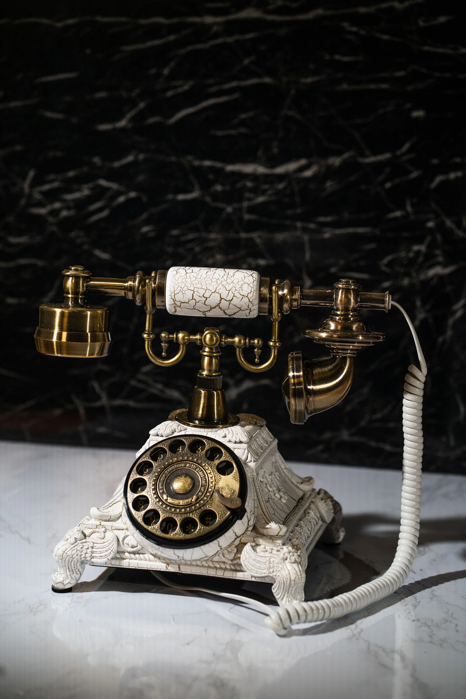 Vintage white and gold rotary telephone displayed on dark surface in dramatic lighting.