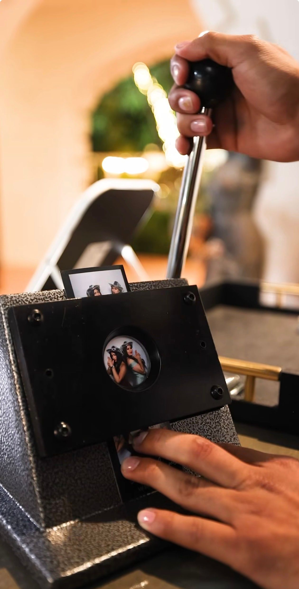 Hands operating a vintage photo booth machine with a lever, displaying small photo strip images inside.