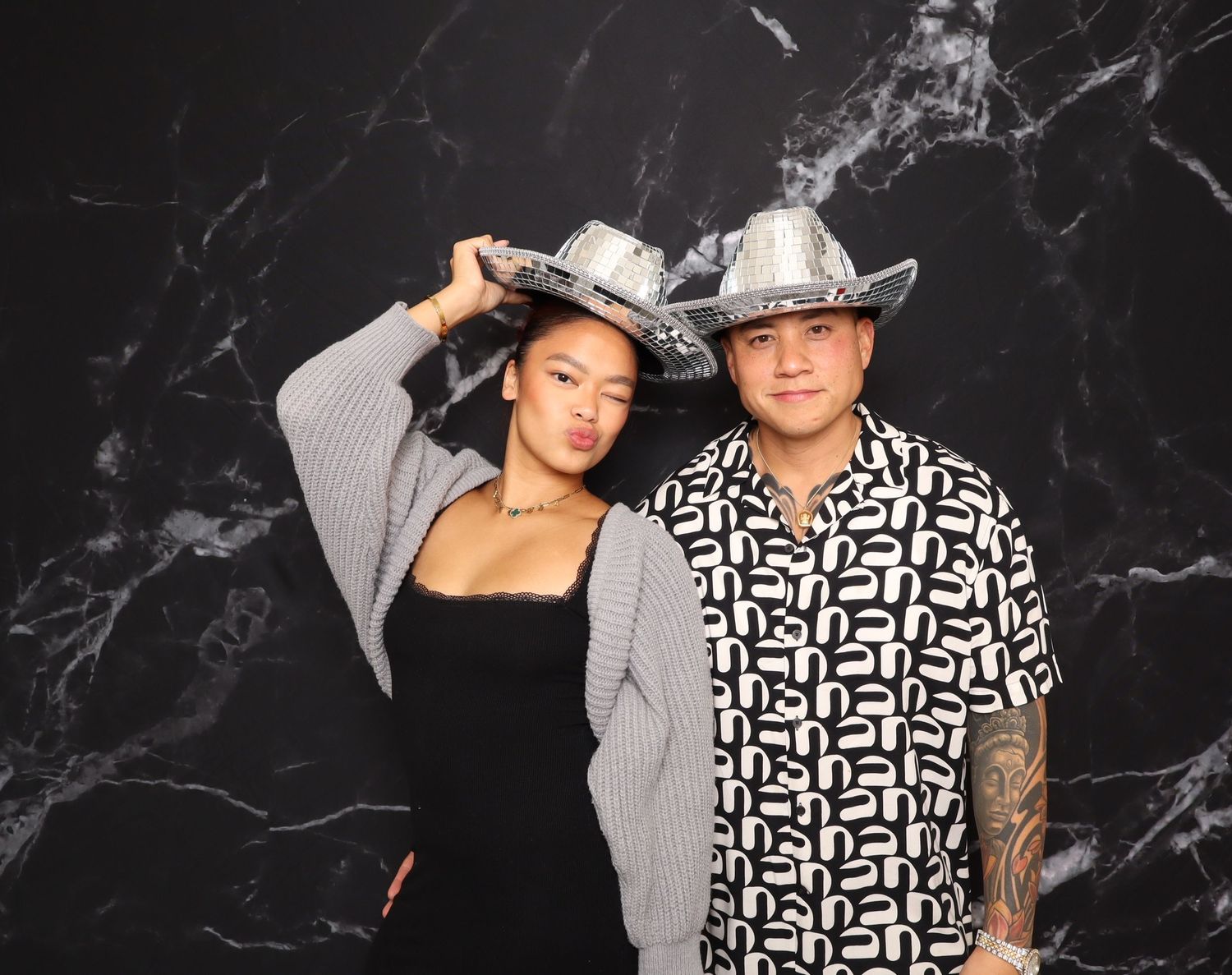 Two people wearing silver cowboy hats pose together against a dark marble background at a photo booth event.