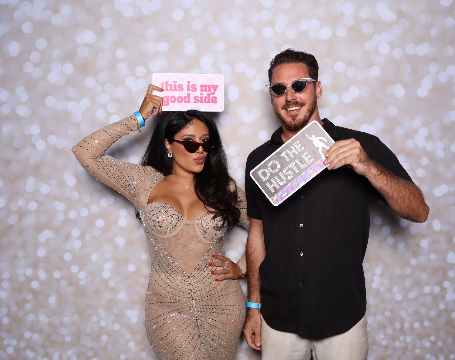 Two people posing at a photo booth holding fun signs, woman in sequin dress and man in striped shirt, bokeh background.