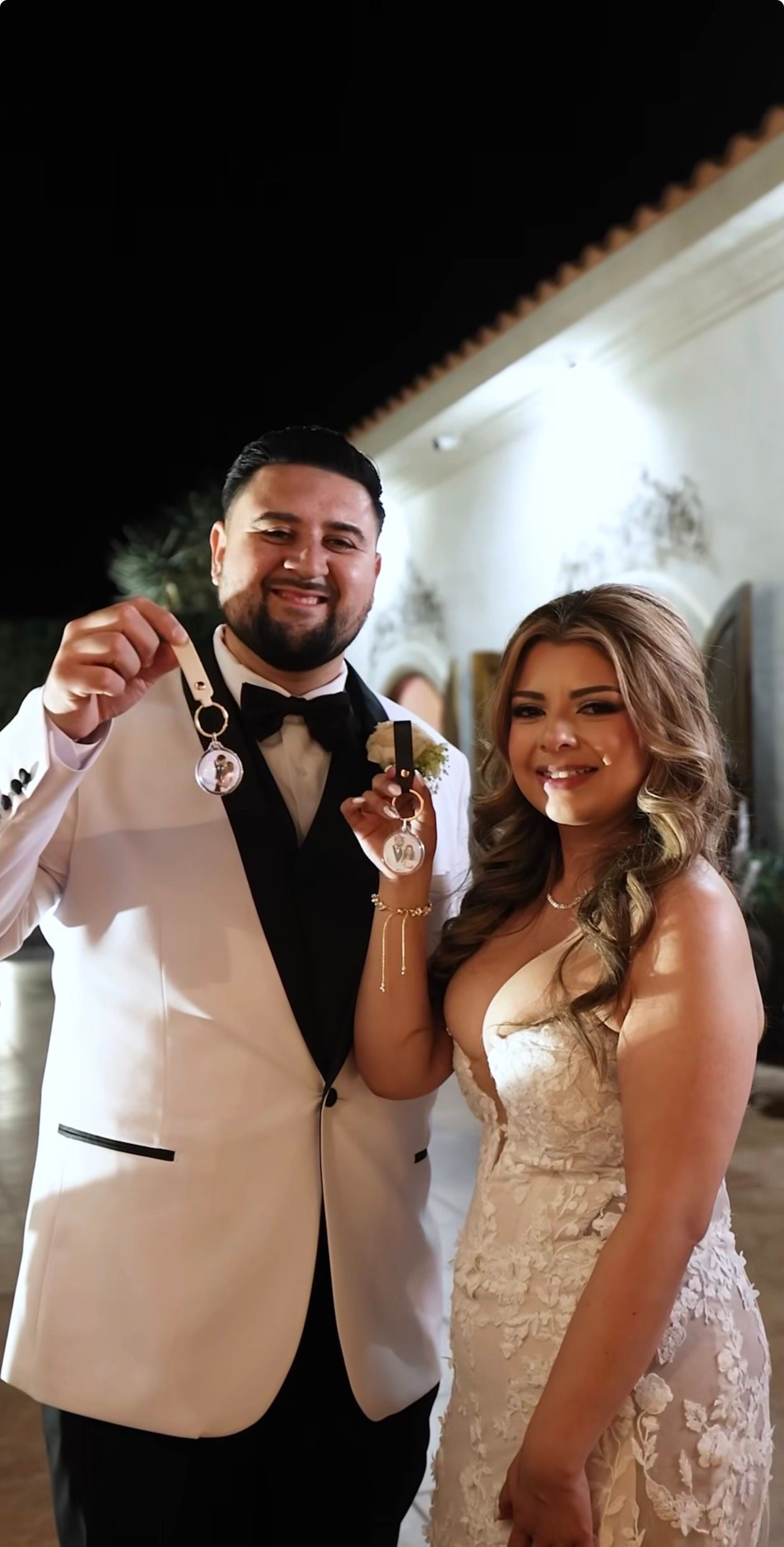 Smiling bride and groom in white tuxedo holding wedding keepsakes outdoors at night.