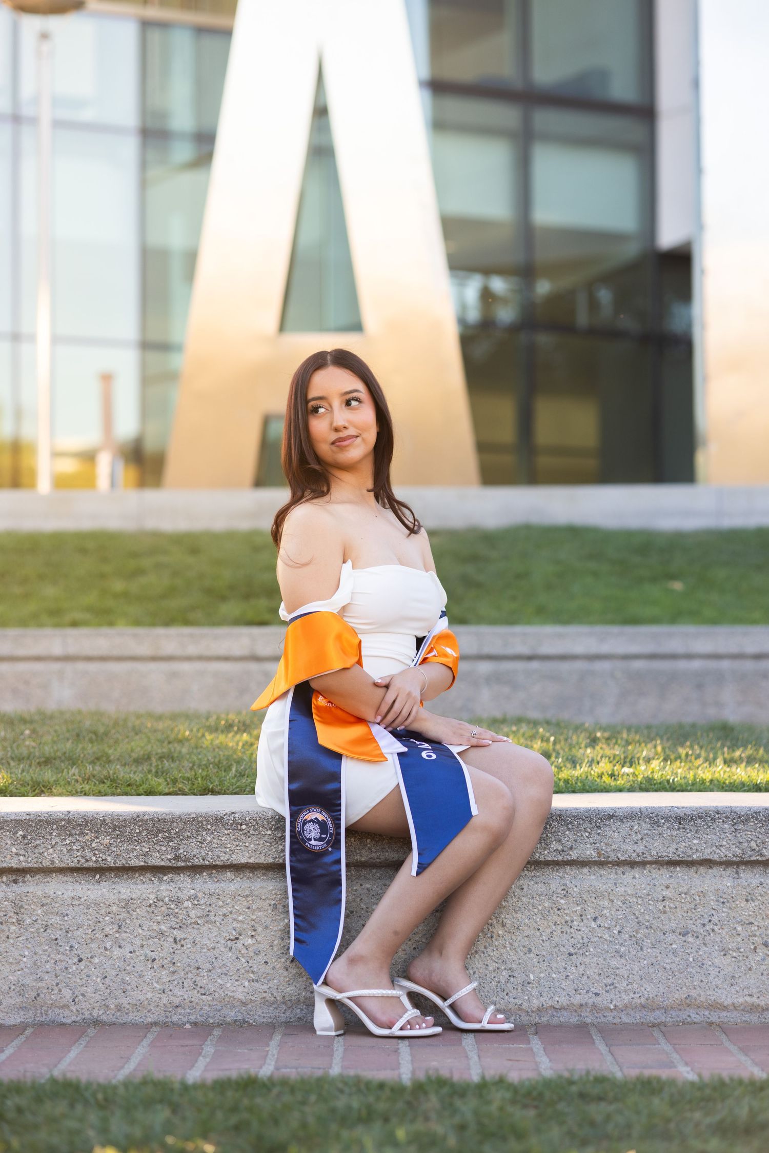 Young woman in white off-shoulder top holding graduation sash, seated on stone steps outside modern university building.