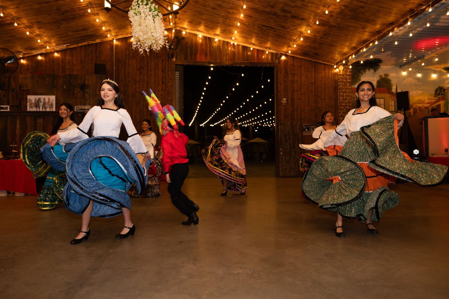 Dancers in colorful full skirts performing in a wooden reception hall with string lights.