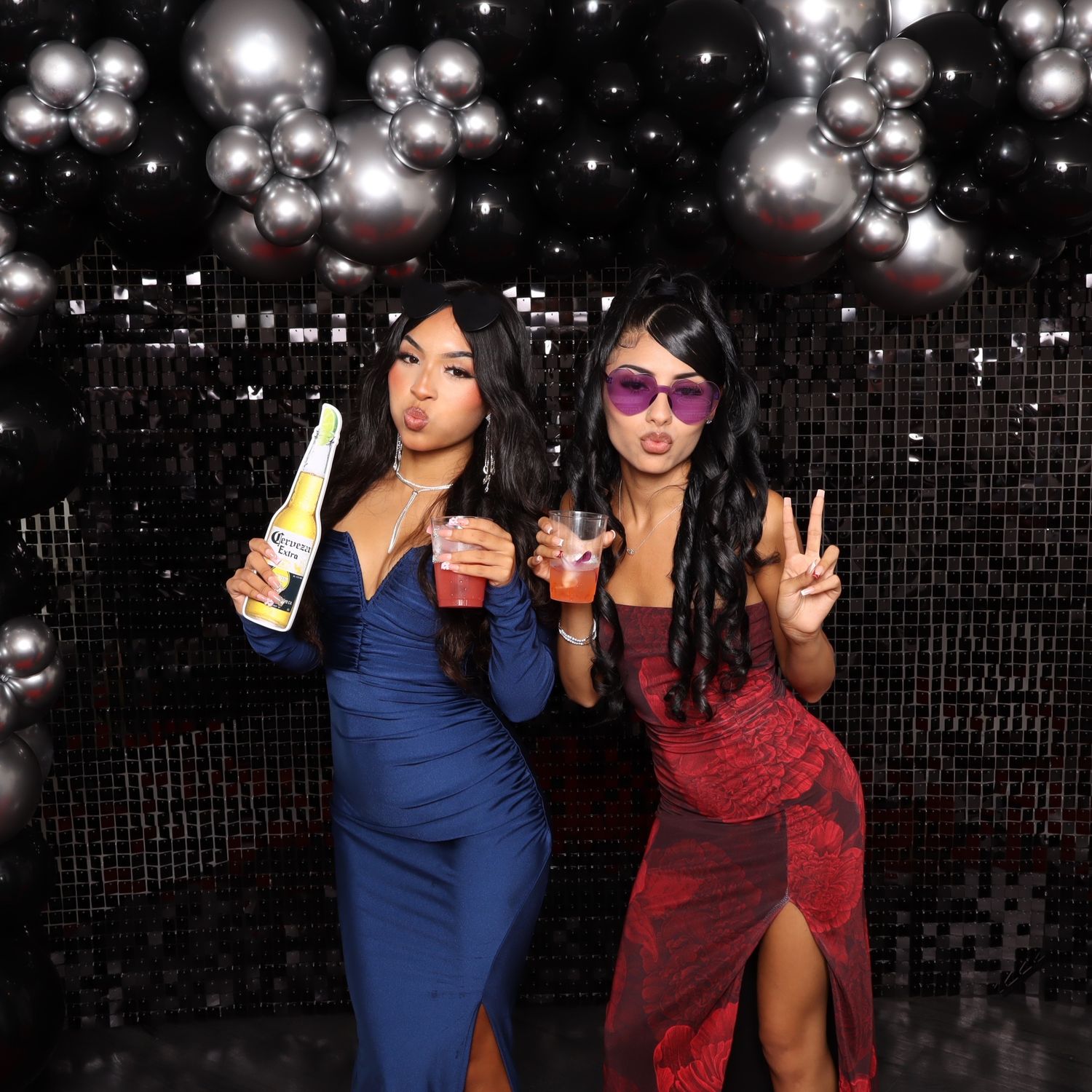 Two women in elegant dresses pose with drinks at a party, surrounded by black balloon decorations.