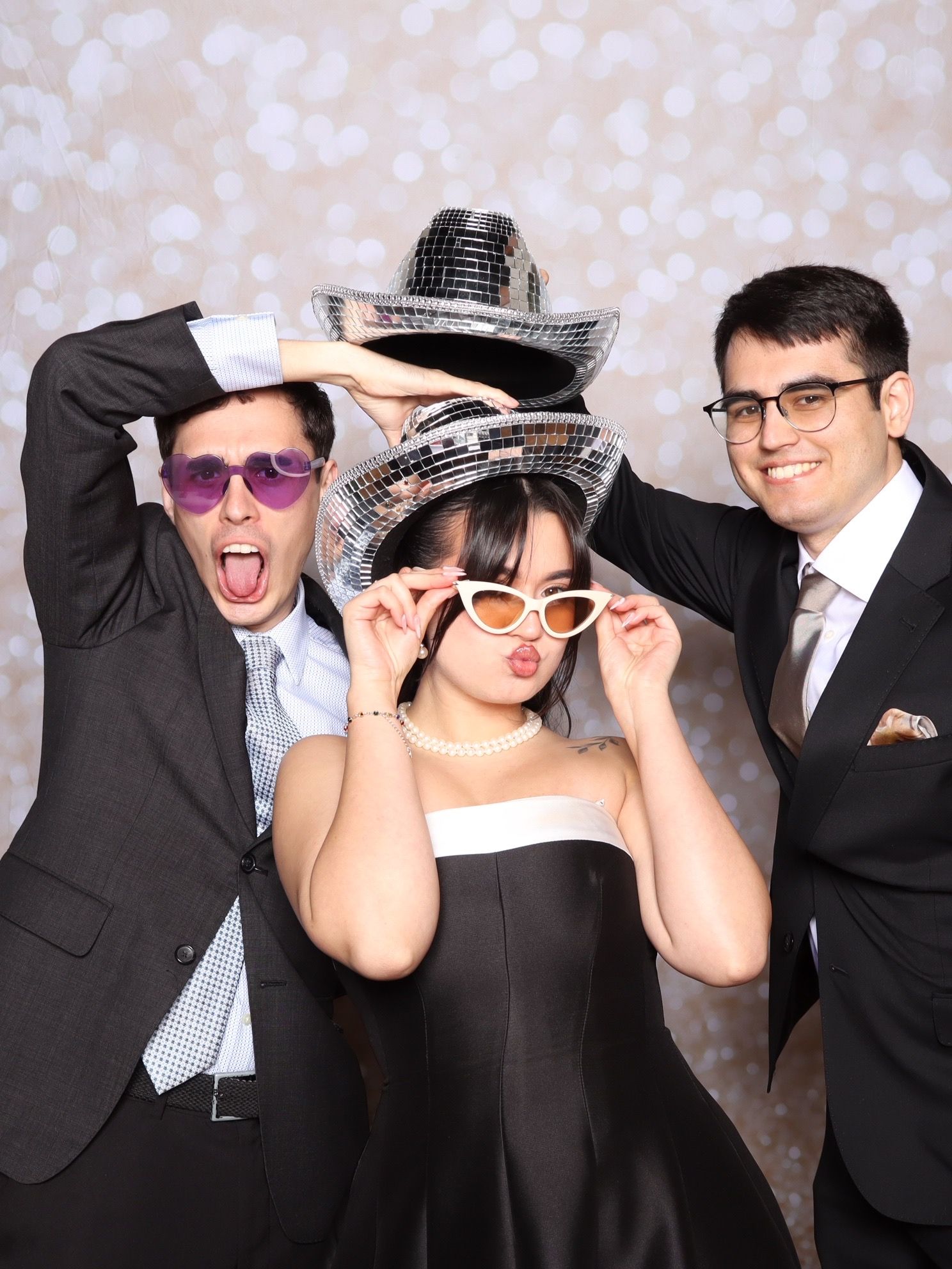 Three friends posing in a photo booth wearing fun accessories including disco ball hats and colorful sunglasses at a formal event.